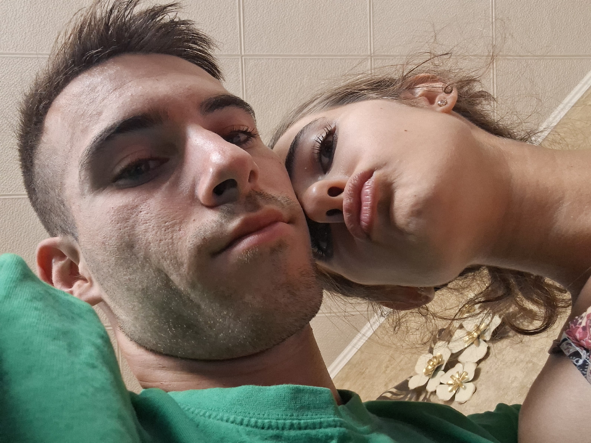 In a close-up, low-angle selfie, two individuals are positioned in the foreground. The upper individual, a male, is seen from the forehead to the chin, with his face tilted downwards. He is wearing a green hooded sweatshirt. Partially obscuring him is the girl's head, who is directly below him. She is looking upwards towards the camera, with her lips puckered in a kiss pose. She has dark makeup on her eyes. Behind them, a tiled wall is visible. To the left of the girl's head, there are decorative floral elements on a textured surface. The lighting suggests an indoor environment. The scene appears to be set in an unknown urban location.