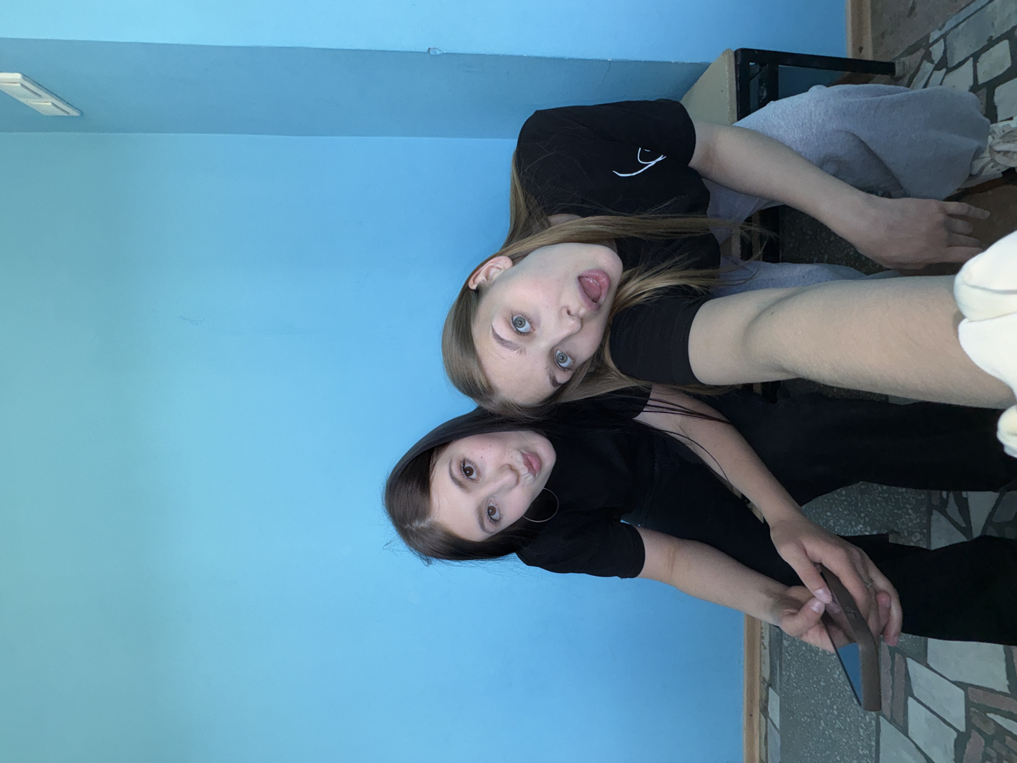 Two young women pose for a selfie in an interior setting with a light blue wall. The woman on top has light brown hair and is sticking out her tongue. She wears a black shirt. The woman below has dark brown hair and is holding a smartphone. She wears a black shirt. The background includes a cabinet, gray sweatpants, and a patterned floor. The location may be in Chaikovsky, Russia.