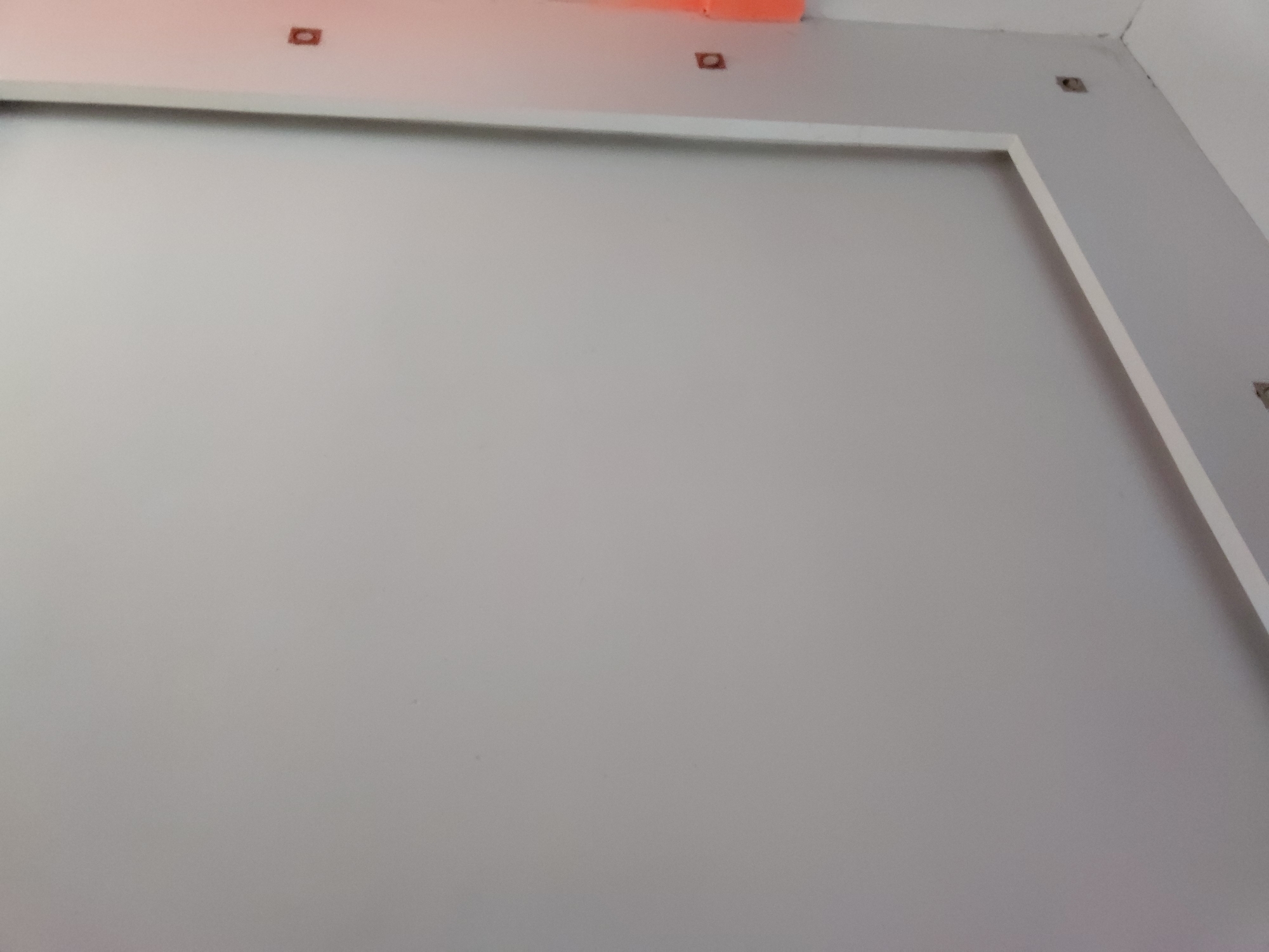 The image displays an interior view of a room, likely undergoing construction or renovation. A large, light gray wall dominates the scene, bordered by a prominent white rectangular frame or trim. Above this frame, a section of the wall or ceiling is visible, painted in a light gray color that matches the main wall, and further up, the ceiling appears white. Near the top edge of the image, an orange or reddish-colored surface, possibly a beam or part of the ceiling structure, adds a contrasting element. Several small, square cutouts or empty sockets are visible near the ceiling line and in the upper right corner, indicating unfinished electrical work or mounting points. The room appears brightly lit, suggesting either interior lighting or ample natural light, though no specific time of day or weather can be determined. There are no people or discernible activities. No text is visible in the image. The scene is neutral and functional, typical of an unadorned or partially completed architectural space. The location context provided is Santa Ana, Venezuela.