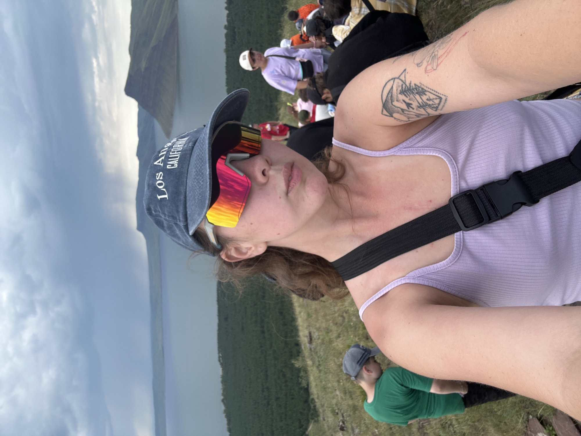 In an outdoor scene likely in the city of Nikolo-Petrovka, Russia, a young woman is taking a selfie. She is wearing a light purple tank top, a blue baseball cap with "Los Angeles California" embroidered on it, and striking, multi-colored reflective sunglasses. She has curly brown hair and a tattoo on her right arm depicting geometric shapes and trees. A black strap from a bag is visible across her chest.
In the background, a body of water, possibly a river or lake, is surrounded by rolling hills covered in dark green forest. The sky is overcast and appears to be threatening rain, with dark clouds dominating the upper portion of the image.
Several other people are scattered across the grassy hillside. To the woman's left, a young person in a green t-shirt and a gray baseball cap is looking down. Further back and to the right, a person in a white cap and purple t-shirt is visible, along with other individuals sitting and standing, some wearing orange and yellow clothing. The overall impression is of a group outing in a natural, somewhat rugged landscape.