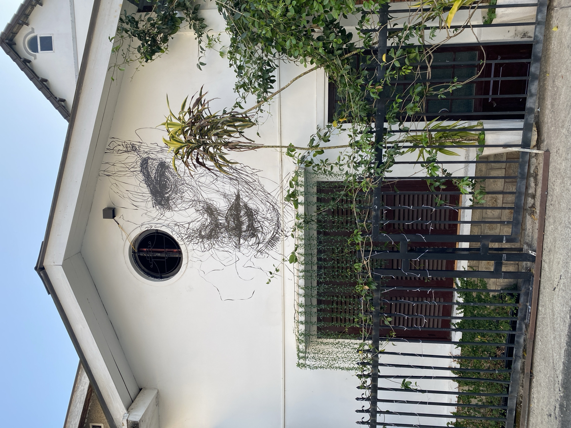 The image shows the exterior of a white, two-story building with a unique face-like design on the wall to the left of center, constructed of dense, dark lines. A round window with a metal frame is situated within the design. Green foliage drapes over the side of the building. The building has two windows with brown shutters and a black metal gate in front. The gate is also adorned with green vines and small string lights. The photograph was taken in Da Lat, Vietnam.