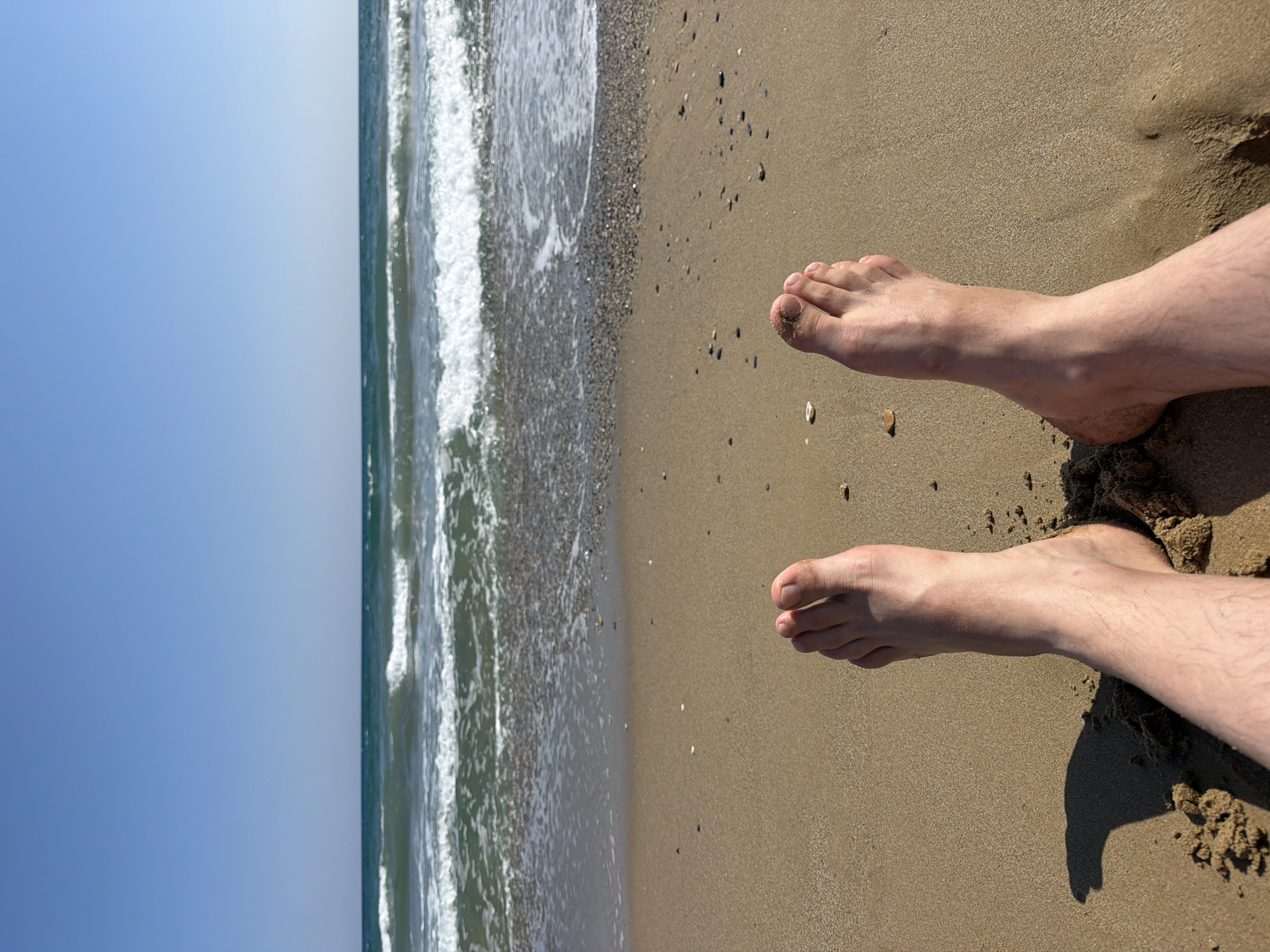 The image shows a first-person perspective of two bare feet resting on a sandy beach. The feet are positioned in the foreground, with the toes pointing upwards and slightly outwards. The sand is a light brown color and appears to be slightly wet in some areas. Behind the feet, the beach continues for a short distance before meeting the water. The ocean is visible in the background, with gentle waves rolling towards the shore. The water is a deep blue-green color, and the sky above is a clear, bright blue. Small pebbles and shells are scattered across the sand near the water's edge. The scene suggests a relaxed moment on a sunny day at the beach, possibly in Georgioupoli, Greece, given the typical clear blue skies and sandy shores characteristic of the region.