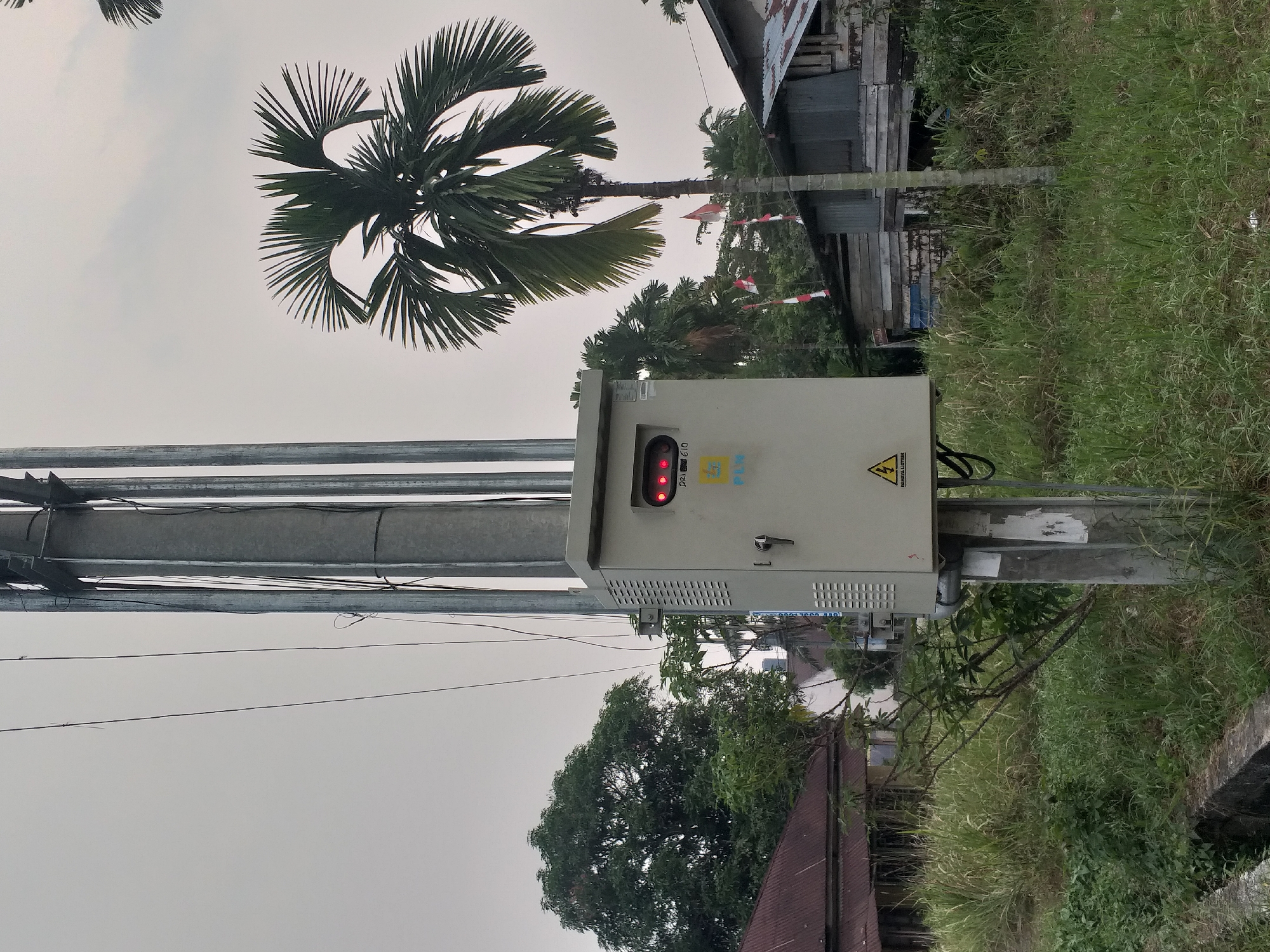 The image captures an outdoor scene in Duri, Indonesia, dominated by a utility pole with an attached electrical box. The utility pole is made of metal and appears to be supporting electrical wires. Mounted on the pole is a cream-colored electrical cabinet, featuring a digital display showing red lights, a ventilation grille, a lock, and a yellow logo with blue text that reads "PLN". A lightning bolt symbol indicating danger is also visible on the cabinet.
Surrounding the pole and cabinet is overgrown green grass and vegetation. In the background, there are several structures. To the left, a building with a reddish, corrugated metal roof is partially visible, nestled amongst lush green trees. To the right of the utility pole, a palm tree with prominent fronds stands tall. Further in the background on the right, two rustic wooden houses with corrugated metal roofs are situated, with Indonesian flags planted between them. The sky is overcast and a pale gray. The scene suggests a rural or semi-urban setting.