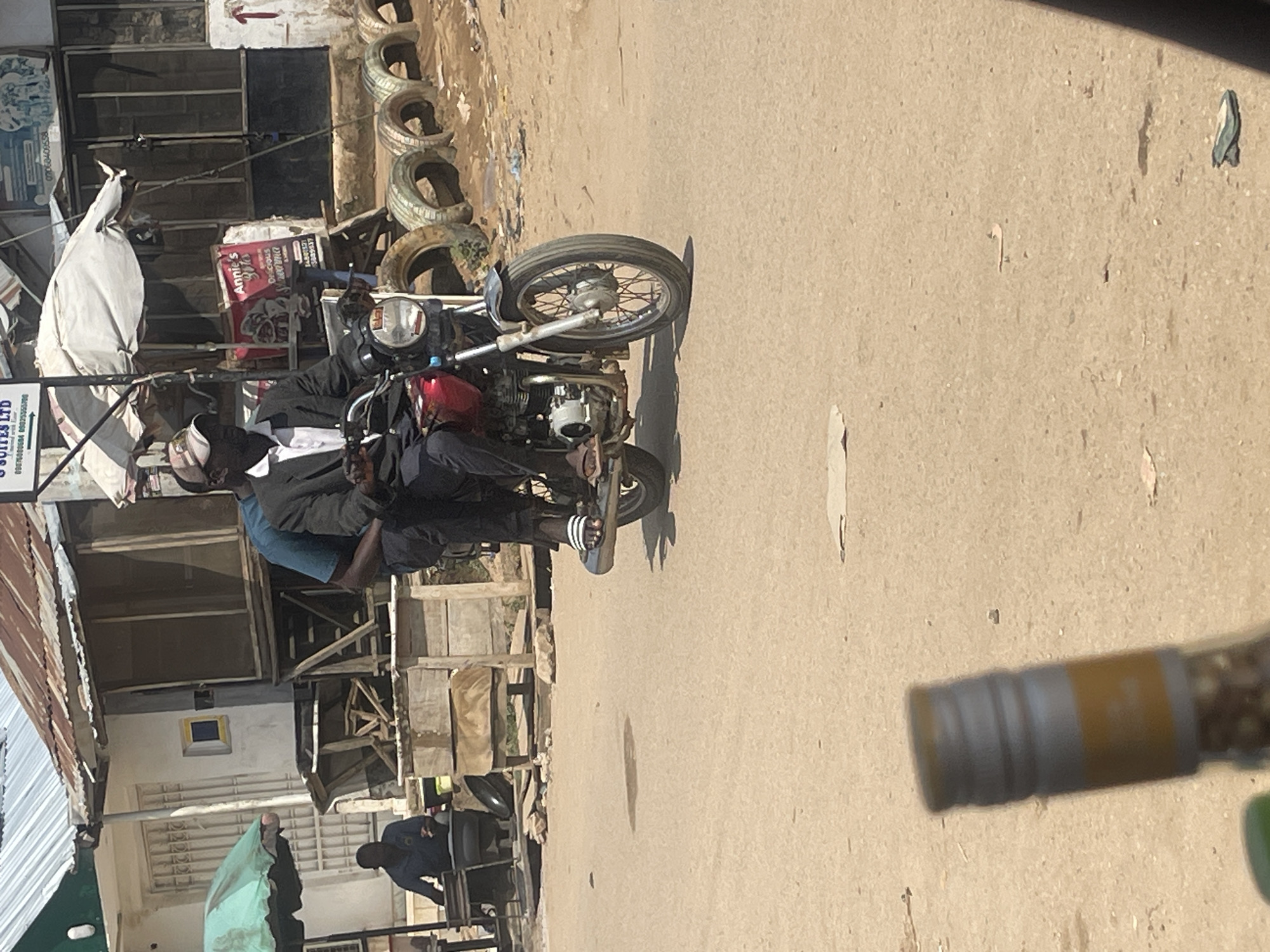 Two men are riding on a motorcycle. The man in front is wearing a dark suit jacket, a white collared shirt, and a baseball cap. He is operating the motorcycle. The second man, wearing a blue shirt, is seated behind him. They are on a paved road in what appears to be a bustling urban environment. In the background, there are buildings with corrugated metal roofs, some with signs, suggesting commercial activity. A person is seated at a stall or shop to the left. The ground is dusty and there are several old tires arranged along the side of the road. The scene is likely in Abuja, Nigeria, given the context of the prompt.