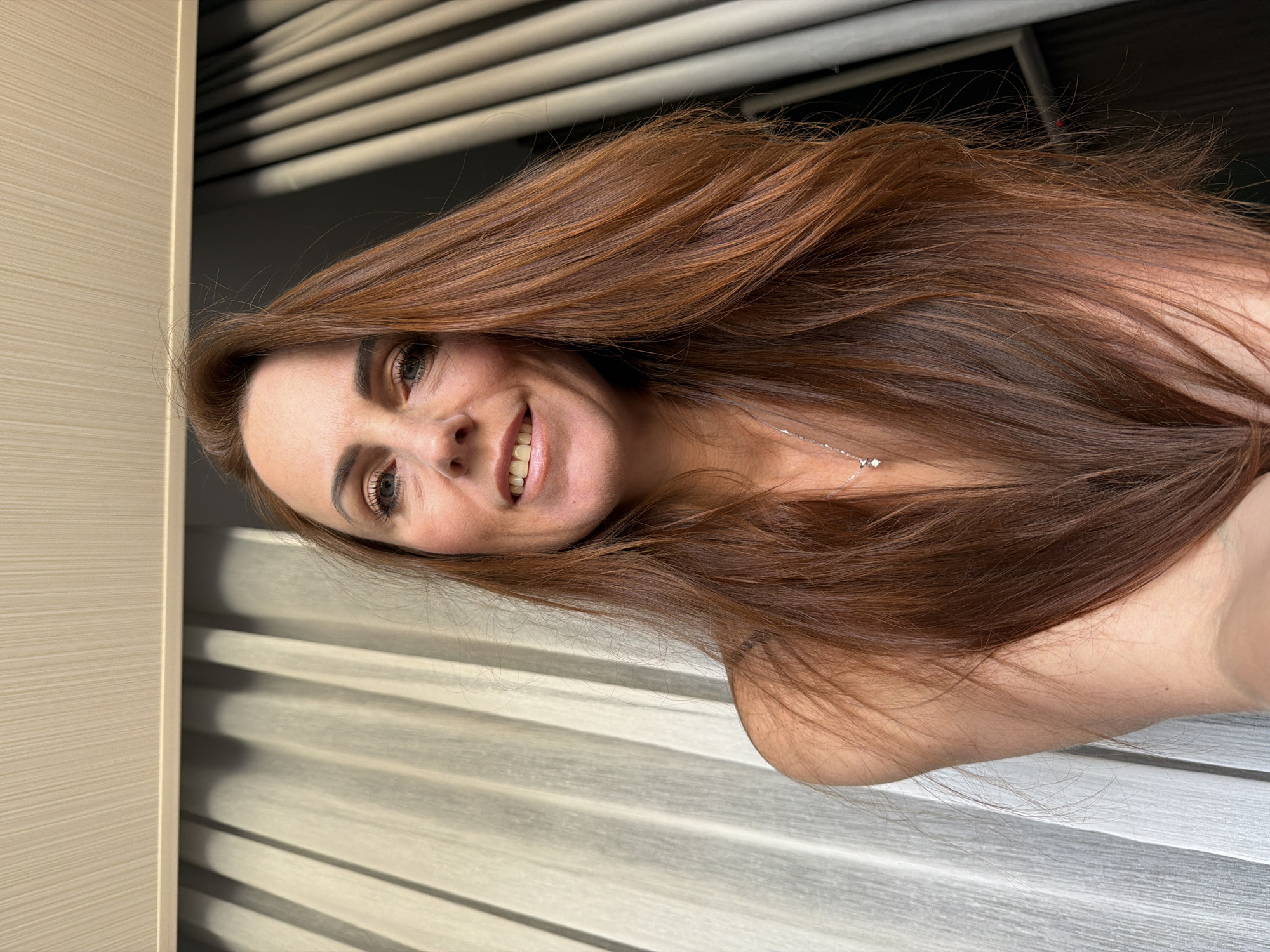 A medium shot, chest-up selfie of a woman with long, reddish-brown hair, is captured from a slightly low angle. She has light skin and is smiling, with her mouth slightly open. Her eyes are green and she is wearing makeup, including eyeliner and mascara. A delicate silver necklace with a small diamond pendant is visible around her neck. The background consists of vertical blinds, with the left side appearing lighter and more translucent than the right side, which is darker and more opaque. The ceiling above the blinds has a textured, ribbed pattern. The lighting suggests it is daytime, possibly indoors near a window. The image does not contain any information about the city of Birobidzhan, Russia.