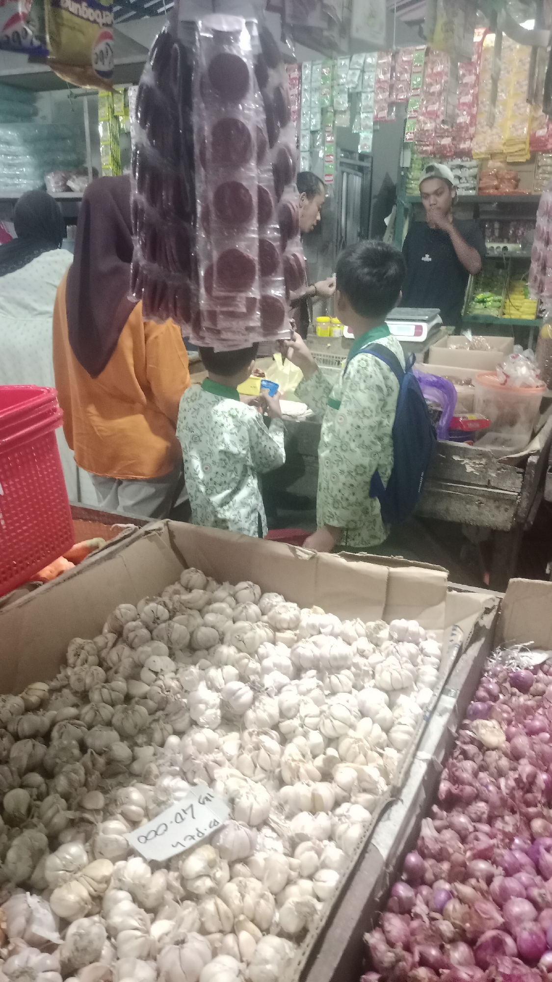 The image depicts a bustling market scene in Karyamandala, Indonesia, likely a wet market or a small grocery store. In the foreground, a large cardboard box overflows with numerous bulbs of garlic, some with price tags. To the right of the garlic, another box contains a pile of red onions. Several individuals are present. A person wearing a black t-shirt and a cap stands behind a counter, seemingly interacting with customers. Two children, one in a light-colored patterned shirt and the other in a similar shirt with a blue backpack, are also visible in the area. In the background, shelves are stocked with various packaged goods and produce, suggesting a diverse range of items available for sale. The overall atmosphere appears to be one of commerce and daily activity within a local Indonesian marketplace.