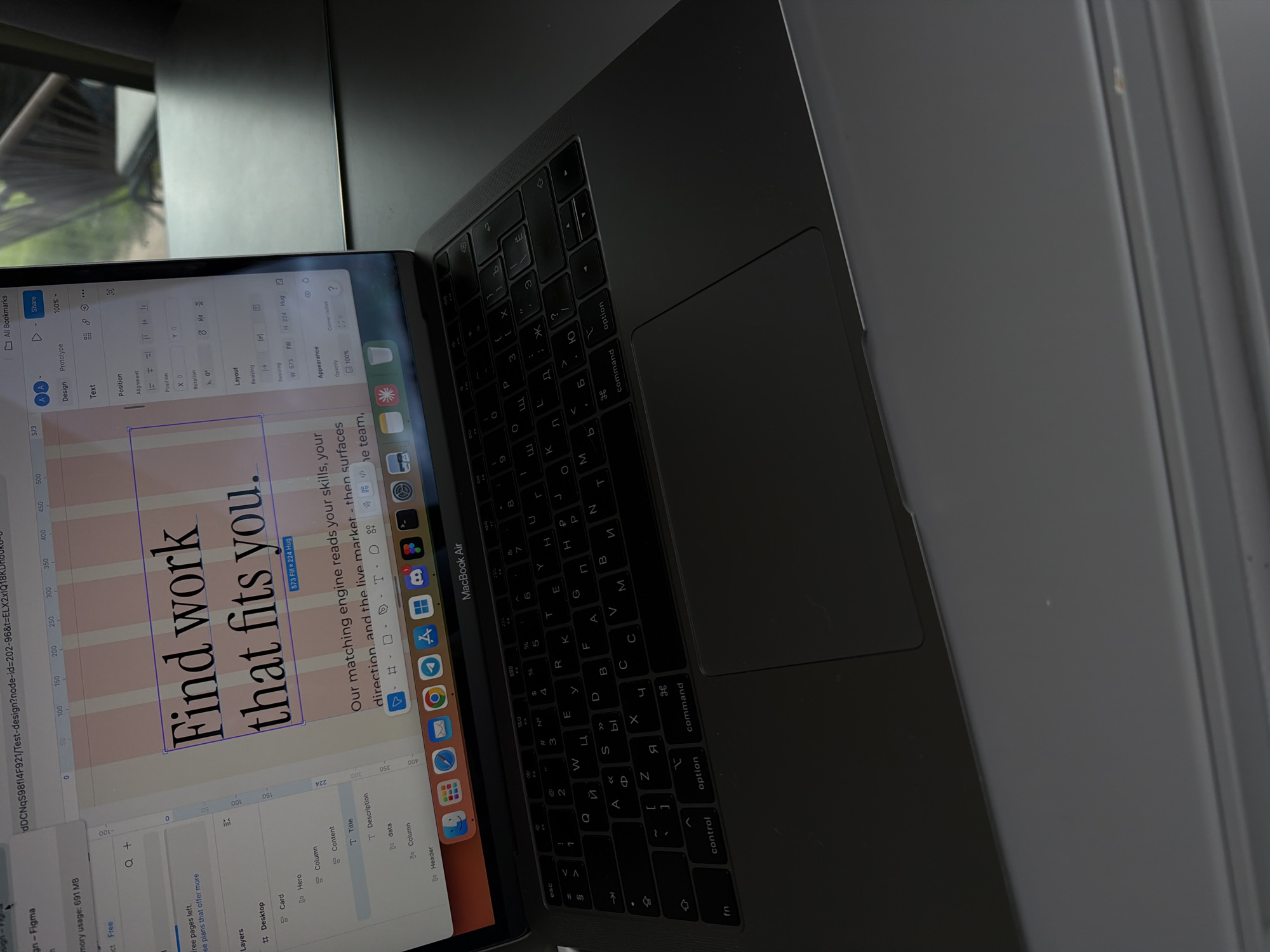 The image shows a MacBook Air laptop open, displaying a design project in Figma. The design features large text reading "Find work that fits you." in a bold font, with pink columns behind the text. Below this is the text: "Our matching engine reads your skills, your direction, and the live market - then surfaces he team." The screen also shows the Figma design interface with various tools and panels. The keyboard of the MacBook Air is visible, with Cyrillic characters on the keys. The laptop is placed on a surface near a window with a view of greenery outside.