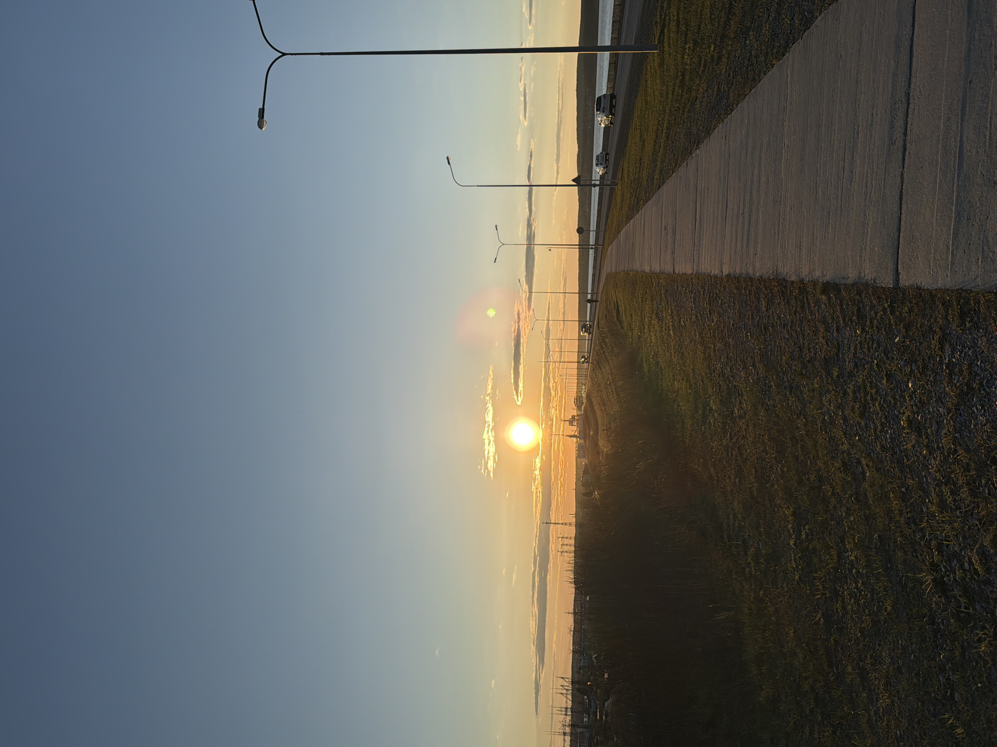 The image shows a sunset scene from Chaikovsky, Russia. The sun is low in the sky, casting a golden light on the horizon. A paved walkway runs along the right side of the image, with grass and some vehicles. A series of streetlights line the walkway. The sky is a gradient of blue at the top, fading into yellow and orange near the horizon.