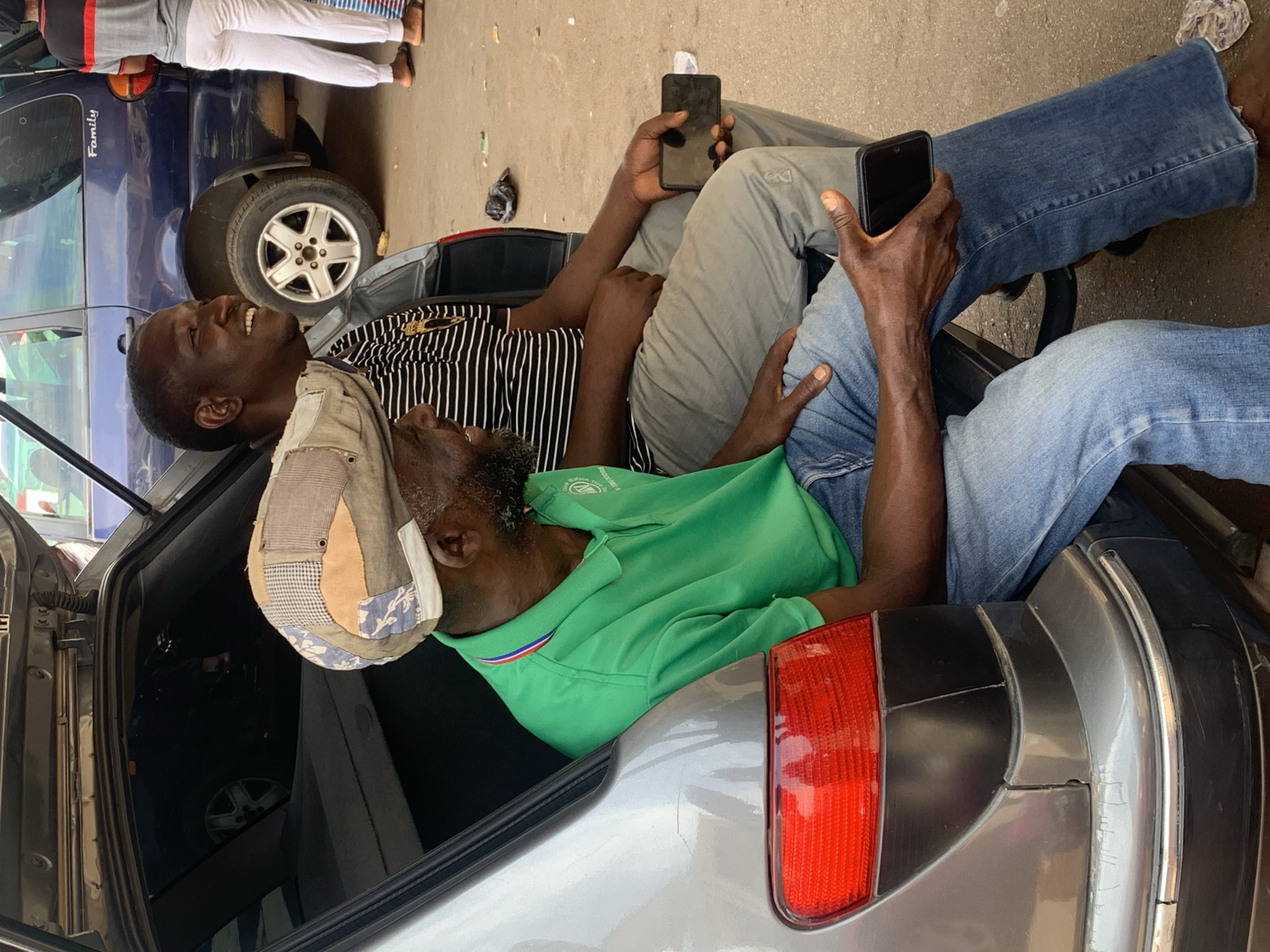 The scene is set on a street in Bukuru, Nigeria. Two adult males are interacting near the open trunk of a silver or light grey sedan. One man is wearing a green collared shirt and a hat; he is reclined in the trunk. The second man wears a black and white striped shirt and is positioned partially in the trunk as well. A third person, dressed in gray pants and shirt, is leaning on the side of a blue car next to the silver sedan and conversing with them. Both men in the trunk are holding cell phones. The street is concrete and shows signs of wear. Another car, a blue vehicle, is parked next to the silver sedan.