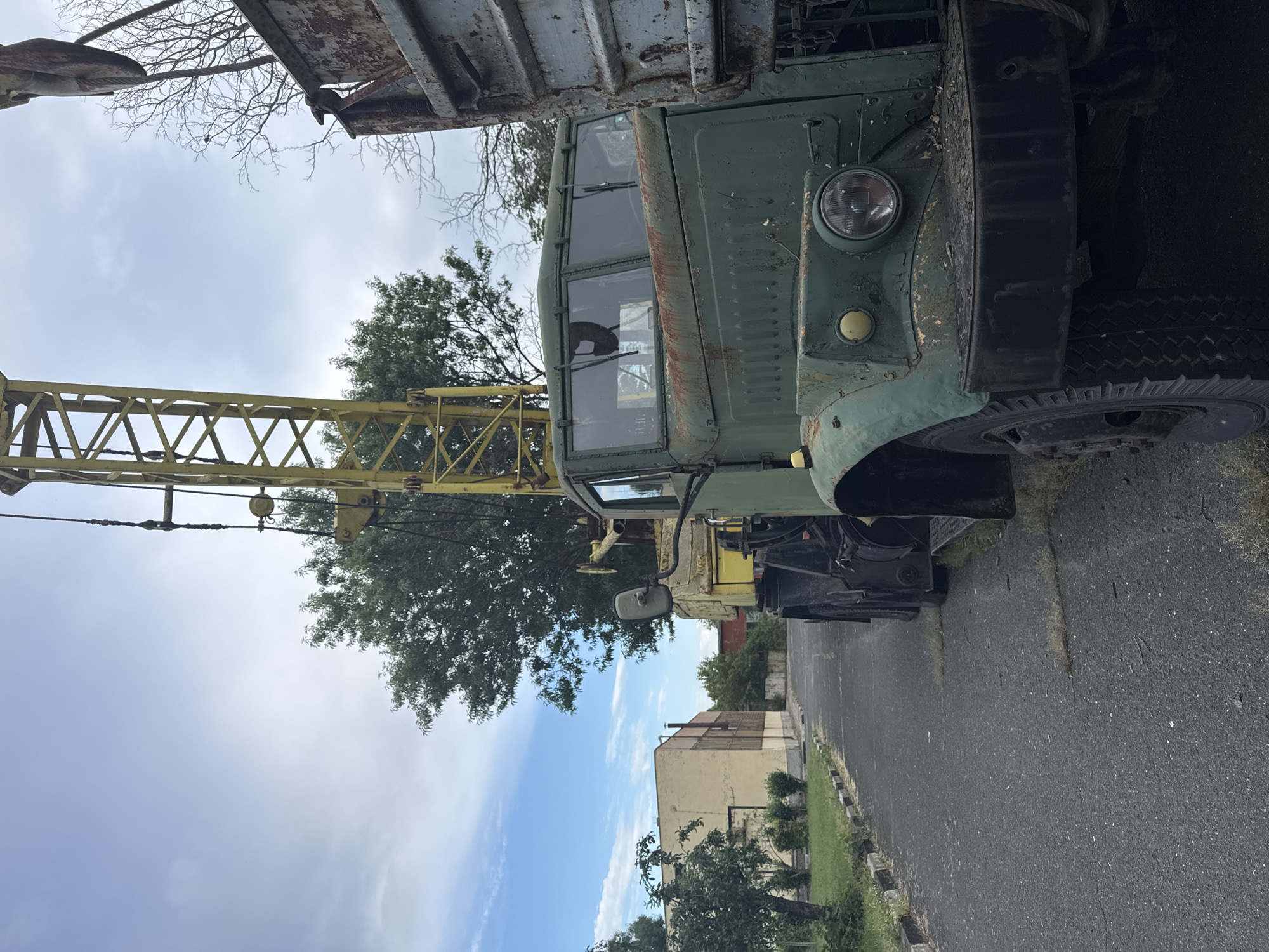 This is a medium shot, taken in portrait orientation, of an old, green military-style truck with a crane attached. The truck appears weathered, with rust visible on its body. It has a classic design with a rounded hood and a large, circular headlight. The crane arm is yellow and extends upwards and to the left of the frame. The scene is set outdoors on a paved surface, possibly a road or parking lot, with a grassy area and some buildings visible in the background. A tree with sparse branches is also present, silhouetted against a cloudy sky. The lighting suggests it might be daytime, but the overcast conditions cast a muted light. The image conveys a sense of abandonment or disuse, as the truck and crane are stationary and appear aged. No people are visible, and there is no text to be read. The overall impression is one of a forgotten piece of machinery in an outdoor, somewhat neglected setting.