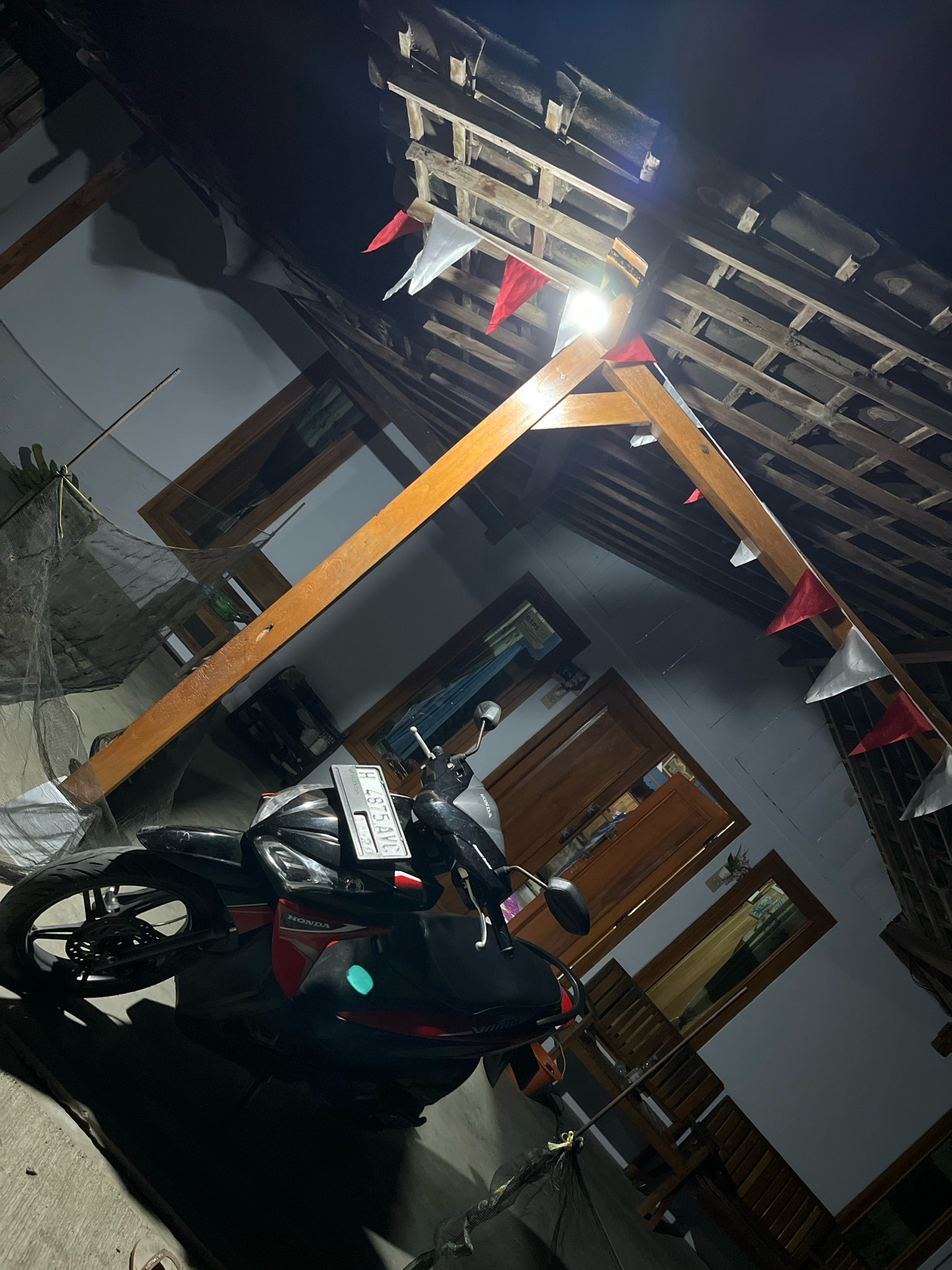 A motorcycle, a black and red Honda Vario, is parked under a wooden-framed structure with a tiled roof. The structure appears to be part of a building, possibly a house, with open doorways visible. Red and white triangular flags are strung along the wooden beams, suggesting a festive occasion. The scene is dimly lit, with a bright light source emanating from the ceiling. A net is draped over a section of the structure on the left. The ground is paved with concrete. The image is taken at night, as indicated by the darkness and artificial lighting. The location is identified as Wonokerto, Indonesia.