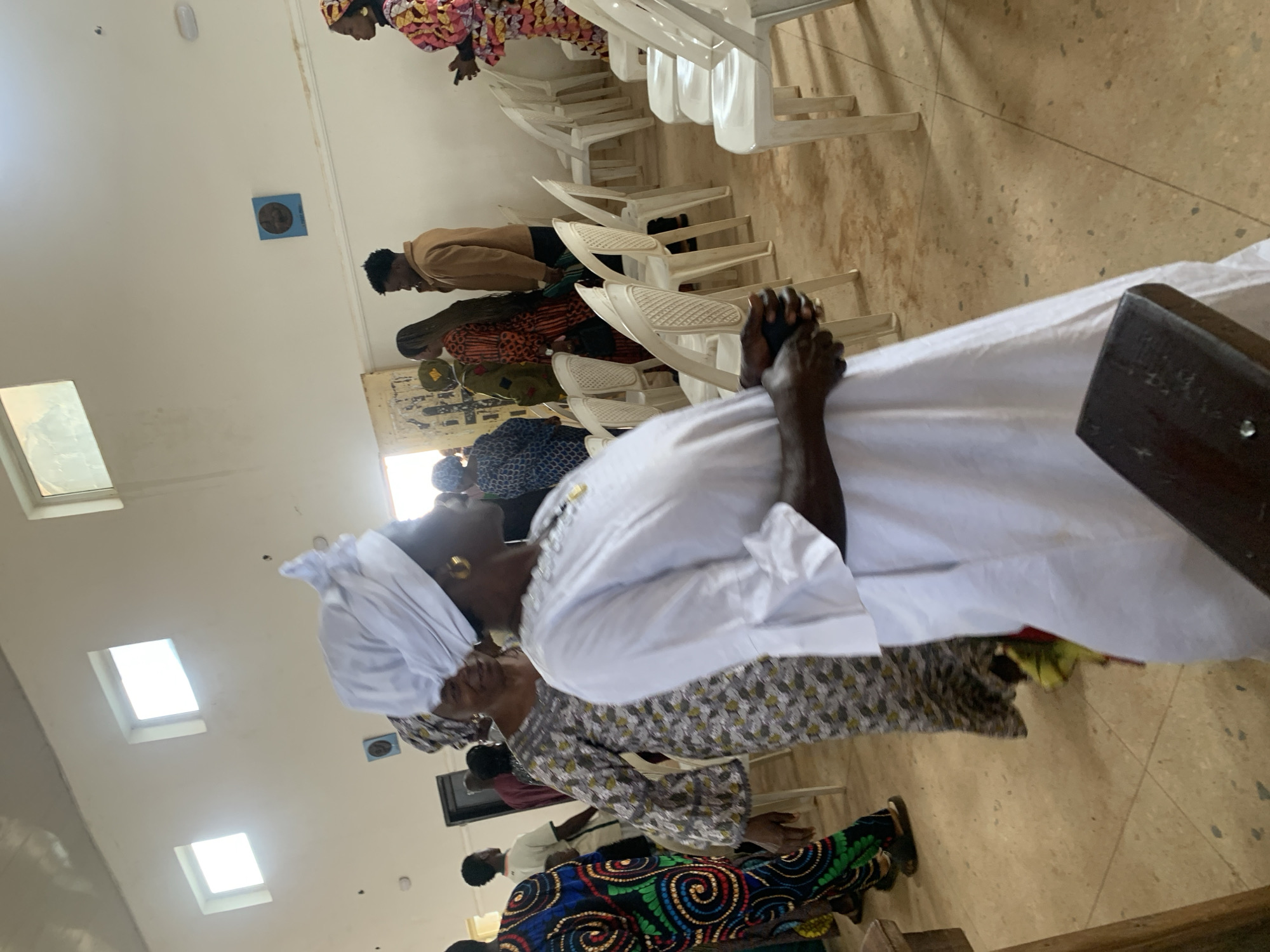 The image depicts an indoor gathering, possibly a religious service or community event, within a brightly lit hall with pale walls. Several people are present, mostly women, dressed in colorful, traditional attire. A prominent figure in the foreground is a woman wearing a white headwrap and a white garment, looking to her right. To her left, another woman in a patterned dress is also visible. In the background, more individuals are seen, some standing, others seated in rows of white plastic chairs. Two square windows are present high on the wall, admitting natural light, and a square blue object is mounted on the wall. The floor is tiled. The overall atmosphere appears to be one of community and social interaction. The specific location in Jos, Nigeria, is not identifiable from the visual cues alone.