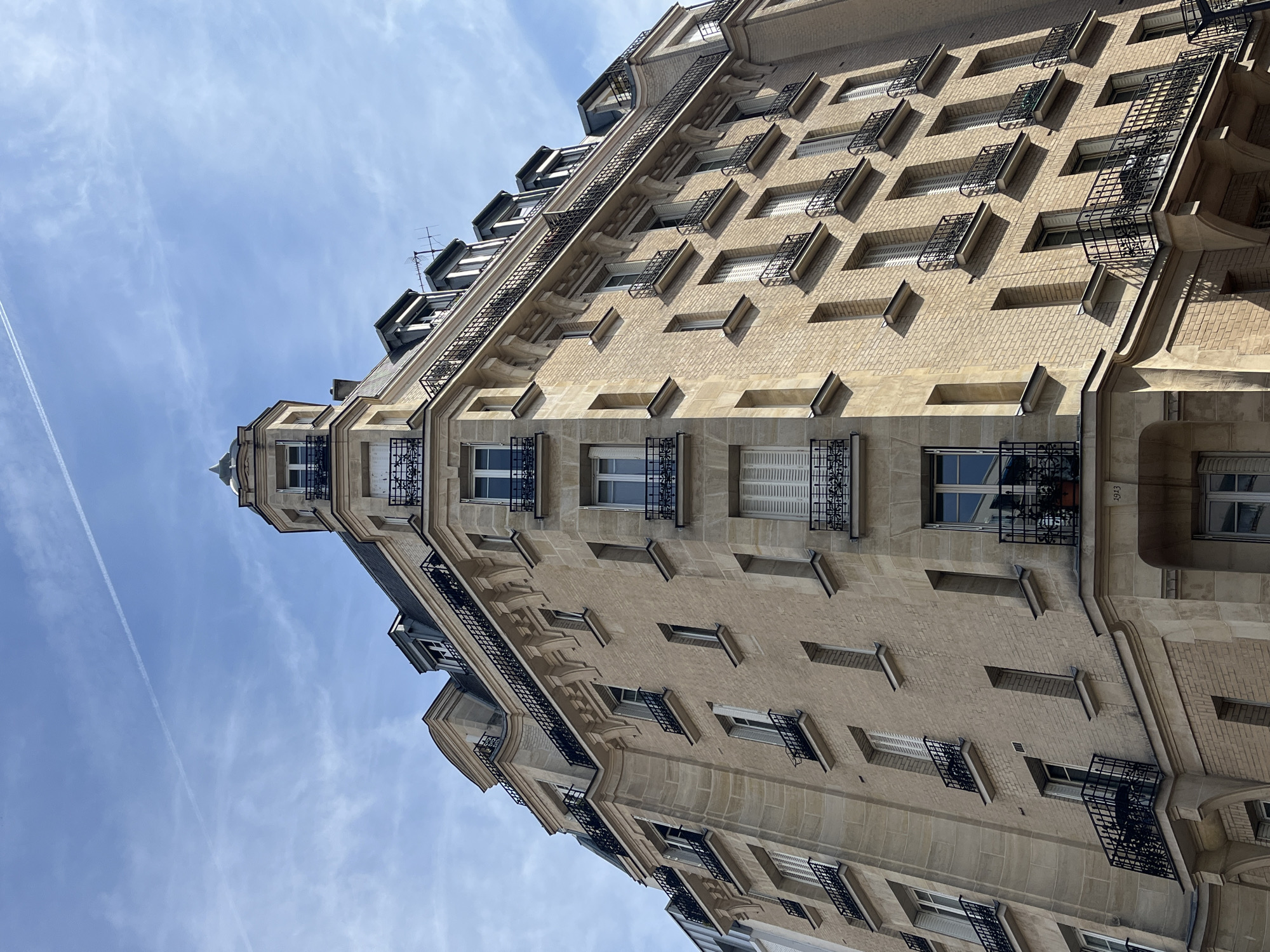 This is a low-angle, full-frame, eye-level exterior shot of a multi-story building with a light brown brick facade and stone accents. The building has multiple windows with black wrought-iron balconies. The windows are a mix of open and closed, with some featuring white shutters. The roofline is visible, with several dormer windows and a small turret with a pointed roof. The sky is blue with some wispy white clouds and a faint contrail. The building appears to be located in Le Kremlin-Bicêtre, France, based on the architectural style. There are no people visible in the image. The overall impression is that of a historic Parisian-style apartment building.