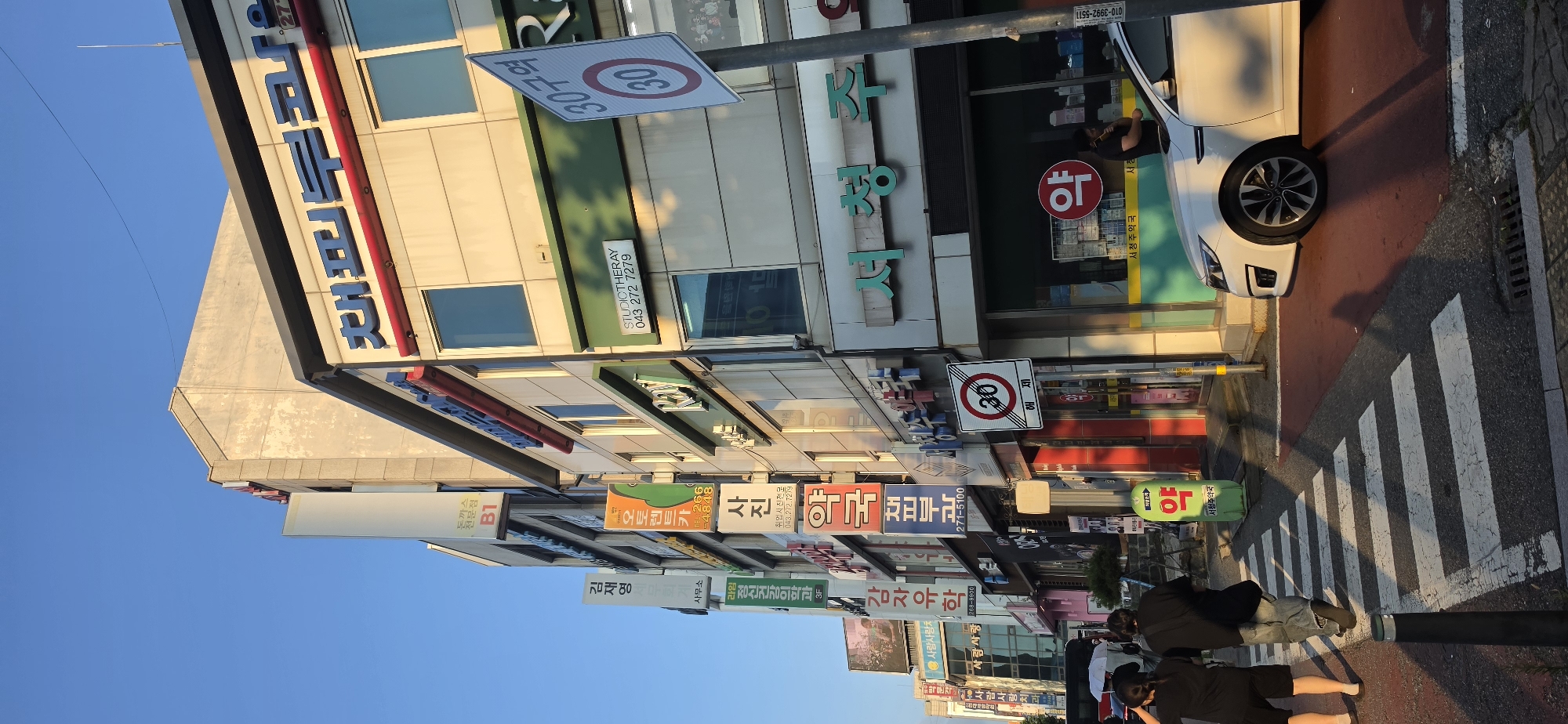 The image depicts a street scene in Cheongju-si, South Korea, on a sunny day with a clear blue sky. The foreground features a crosswalk with white stripes on a red asphalt surface, and two individuals are walking across it from left to right. The person on the left is wearing black shorts and a black t-shirt, while the person on the right is wearing blue jeans and a black t-shirt. A white car is parked to the right, partially obscuring the street.
The midground and background are dominated by a row of multi-story buildings with various storefronts and signage. The buildings have a mix of light-colored facades and glass windows. Prominent signs are visible, including Korean characters and some English text. One sign displays "STUDIOTHERAY 043 272 7279". There is also a traffic sign indicating "30구역" (Zone 30) with a circular speed limit sign showing "30". Another sign with a red circle and the character "약" (medicine) is visible. A bright green, inflatable structure with the same "약" character stands near the sidewalk. A street light pole is positioned to the right of the frame. The overall impression is of a commercial street in an urban environment.