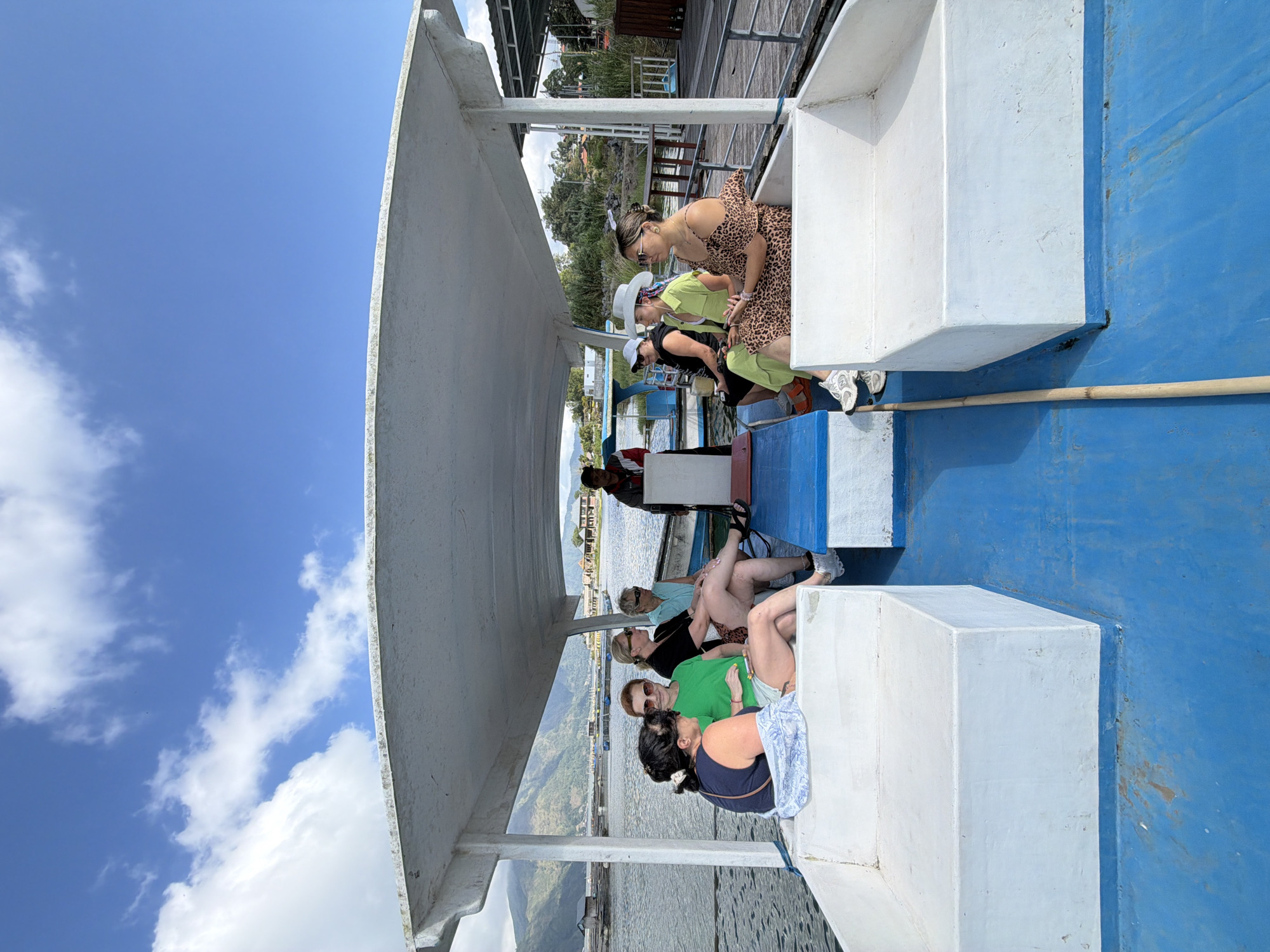 This is a horizontal, eye-level, full shot of people on a boat on a lake in Kintamani, Indonesia. In the foreground, the bright blue deck of the boat takes up the bottom half of the image. White, built-in seating lines the edges of the boat. Several people, a mix of genders, are seated in the white seating areas. On the left, two women are talking to each other, one in a blue shirt and the other in a green shirt. Further back, three more women are seated, one in a black shirt and sunglasses, another in a light blue shirt, and a third woman in a green shirt and white hat. On the right side of the boat, a woman in a leopard print dress is seated, and a man wearing a hat and vest is standing near the back, appearing to be the boat's pilot. The boat is covered by a white awning. In the background, a body of water with some floating structures is visible. Beyond the water, a lush, green landscape with mountains under a bright blue sky with white clouds completes the scene.