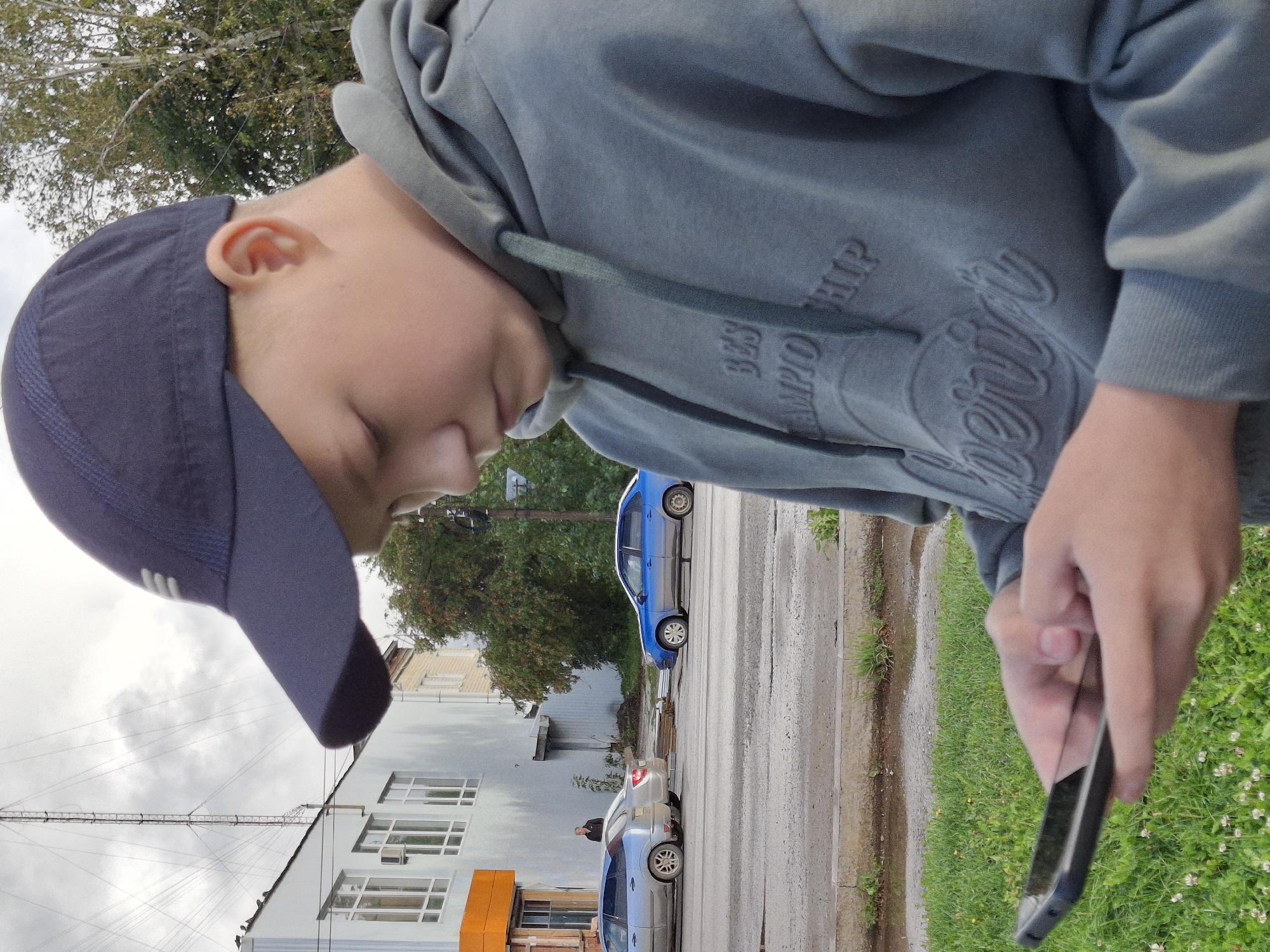 A young boy wearing a dark blue baseball cap and a grey hooded sweatshirt is looking down at a smartphone he holds in his hands. He is standing on a grassy area next to a street in the city of Elovo, Russia. In the background, there is a light blue car and a silver car parked on the street, as well as a building with orange accents. A tall tower and trees are also visible in the background. The sky is overcast.