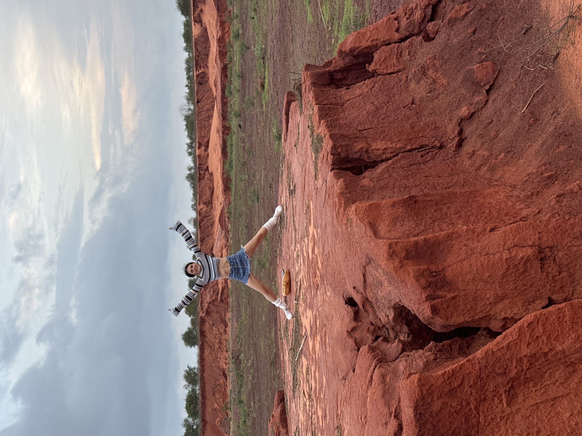 A young woman is captured in a playful pose amidst the distinctive red geological formations of Mui Ne, Vietnam. She stands with her arms outstretched and legs apart, wearing a striped long-sleeved crop top, denim shorts, white socks, and white sneakers. A small, light-brown, gourd-like object rests on the red ground near her feet.
The scene showcases a landscape characterized by deep red-brown, eroded earth and textured rock formations, reminiscent of sand dunes or canyons. In the background, sparse green vegetation and a line of trees delineate the horizon. The sky is overcast with diffused light, indicating a cloudy day rather than harsh direct sunlight. The woman appears joyful and engaged with her surroundings. The image itself is oriented vertically, causing the horizontal landscape to appear rotated ninety degrees. No text is visible in the photograph.