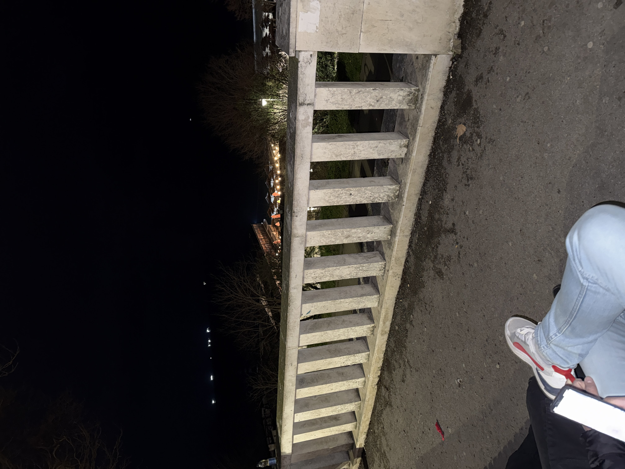 A person stands on the sidewalk next to a concrete bridge. The person is wearing jeans and white sneakers with red accents. The bridge has a railing made of evenly spaced concrete posts. Beyond the bridge, bare trees are visible. In the distance, there are lights that suggest a building or structure with decorative lighting. The location is most likely Varna, Bulgaria, as it's a city known for its architecture and bridges. The scene is captured at night, with a clear distinction between the lit areas and the dark sky.