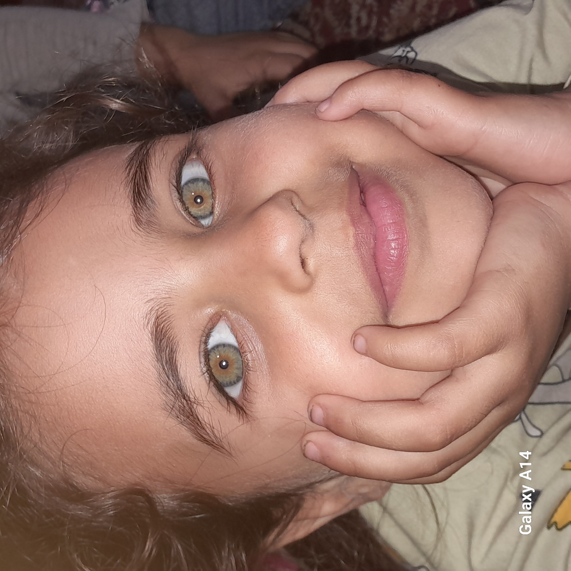 A young girl with curly brown hair and striking hazel eyes is lying down, facing the camera. Her hands are gently framing her face. She has fair skin and pink lips, and her expression is soft and slightly smiling. The background is blurry, showing parts of a patterned fabric. The lower right corner of the image has "Galaxy A14" printed on it. The location is unknown.