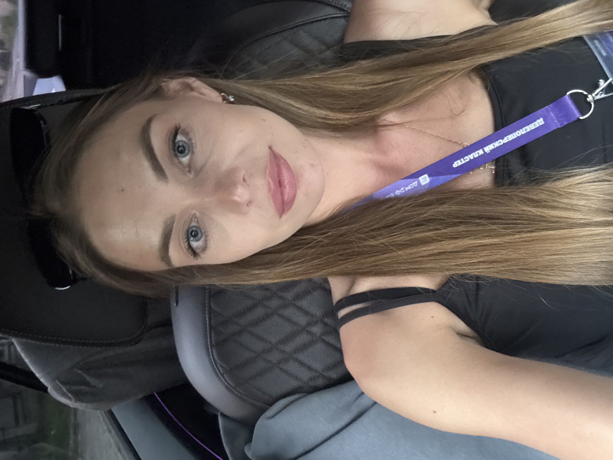 A medium shot captures a woman from the chest up, seated in the backseat of a car. She has long, straight brown hair that falls over her shoulders. Her skin is fair, and she has light blue eyes and a light pink lipstick. She wears a black tank top with thin straps and a purple lanyard around her neck, from which a lanyard clip is visible. Sunglasses rest on top of her head. The background shows the quilted leather interior of the car, with dark gray seats and a hint of purple ambient lighting. A gray blanket is draped over the seat to her left. The image appears to be taken during daylight, with the lighting suggesting an outdoor setting, possibly in the city of Estosadok, Russia.