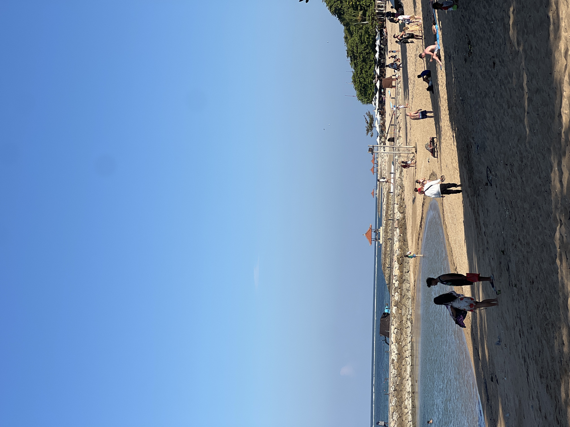 The image depicts a sandy beach in Denpasar, Indonesia, under a clear blue sky. In the foreground, two individuals, a woman in a floral dress and a man in a dark t-shirt, stand facing away from the camera. The beach is populated with several other people, some sunbathing on loungers, others walking or standing in groups. A gentle wave laps at the shore where a stone breakwater extends into the water. Further down the beach, numerous white umbrellas provide shade, and a few small gazebos with orange roofs are visible. Lush green trees line the edge of the beach in the background. The scene captures a relaxed atmosphere with various individuals enjoying the coastal environment.