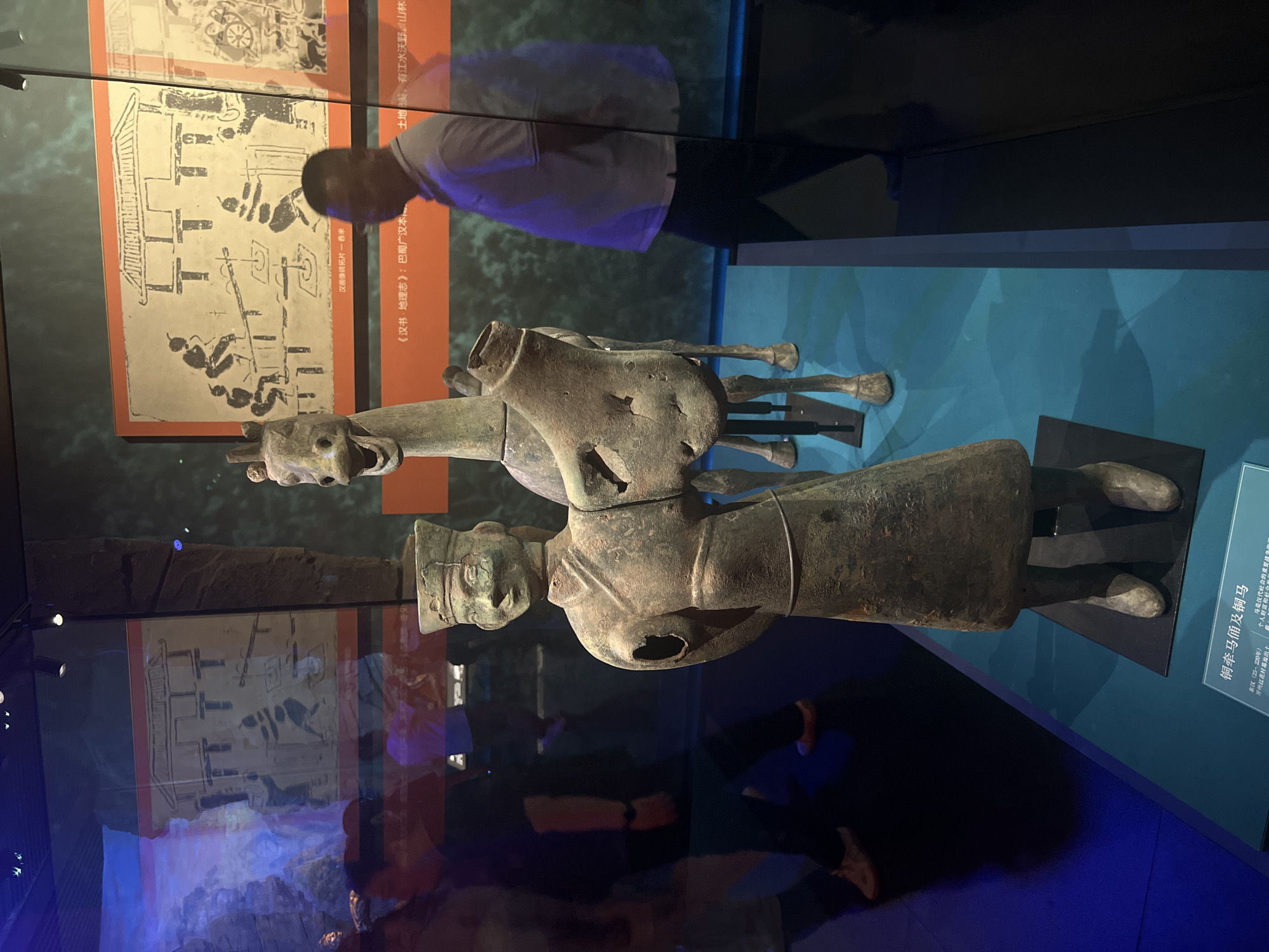 The image depicts a museum exhibit featuring ancient Chinese bronze artifacts, specifically a bronze figurine of a person holding a chariot part and a bronze horse. The figures are displayed on a blue platform. In the background, there are large panels with ancient Chinese artwork depicting scenes of daily life or historical events. Several blurred figures of museum visitors are visible, reflected in the glass display case. One man in a white polo shirt and dark pants is standing to the right of the exhibit. The text at the bottom of the image, though partially obscured, appears to be a label for the exhibit, likely identifying the artifacts and their historical context, mentioning "Yuzhong District, China". The lighting in the exhibit is dim, with spotlights illuminating the artifacts. The overall environment suggests an indoor museum setting, possibly within the Yuzhong District of China.