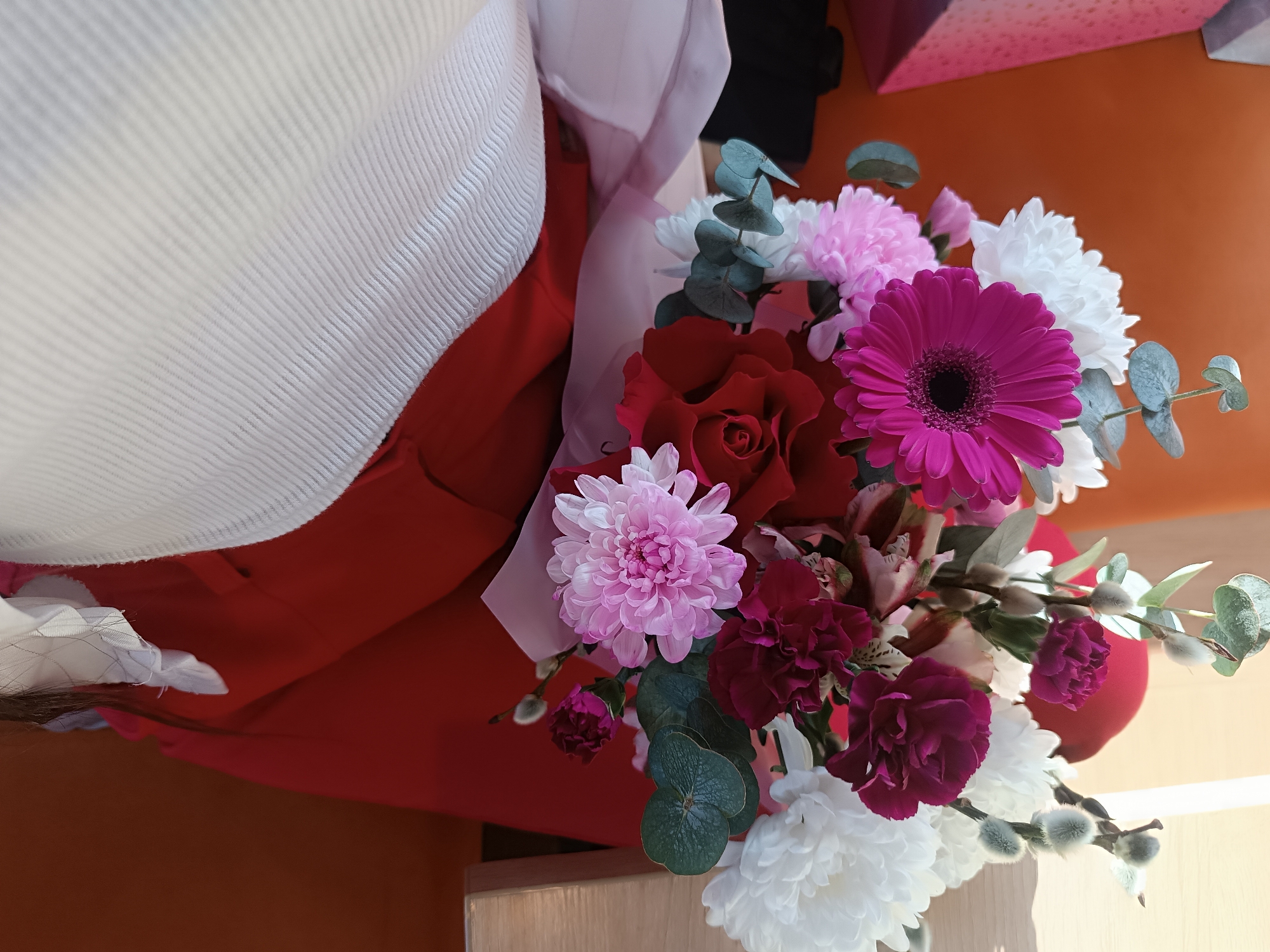 A close-up, angled photograph captures a mixed floral bouquet being held indoors in Chaikovsky, Russia. The bouquet features a prominent magenta gerbera daisy, several red roses, numerous white chrysanthemums, several pink chrysanthemums, multiple dark magenta carnations, light-colored alstroemeria, green eucalyptus foliage, and pussy willow branches with fluffy catkins. The flowers are wrapped in a translucent light pink material and secured with a red or dark pink base. A person's attire is partially visible on the left side of the frame, consisting of a white ribbed garment and a red garment, with a glimpse of dark hair. The background includes an orange-brown wall or surface, a partially visible pink patterned object in the upper right, and a light-toned wooden surface at the bottom.