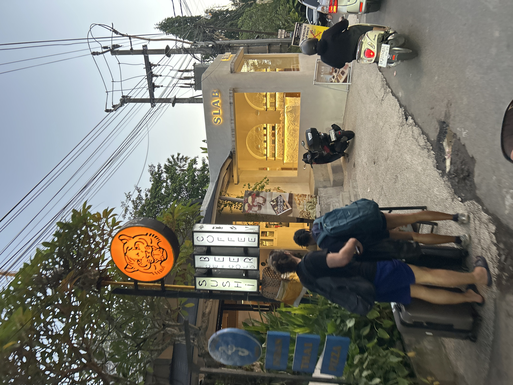 In Ubud District, Indonesia, at dusk, a street scene unfolds with a focus on a brightly lit establishment called "SLAB" which appears to be a shop selling baked goods or confectioneries, as indicated by the display of items in its large glass windows and a sign that reads "HANDCRAFTED CHOCOLATES GELATO". Adjacent to SLAB, a neon sign in a circular orange casing depicts a tiger's face and illuminates the surrounding area. Below this, three vertical illuminated signs read "SUSHI", "BEER", and "COFFEE". A man and a woman, both carrying backpacks, stand with two large suitcases on the sidewalk in front of the shop. One person is wearing a black t-shirt and blue shorts, while the other is wearing a black t-shirt and shorts. A black scooter is parked in front of SLAB. Further down the street, a person on a scooter wearing a black jacket and helmet rides past. The street is paved and shows signs of wear and tear, with a large pothole visible. Overhead, a complex network of electrical wires and utility poles dominate the sky. Several other signs are visible, including a blue circular sign and blue rectangular signs indicating "PIZZA" and "THAI". The overall atmosphere suggests a busy urban environment with a mix of commercial activity and local life.