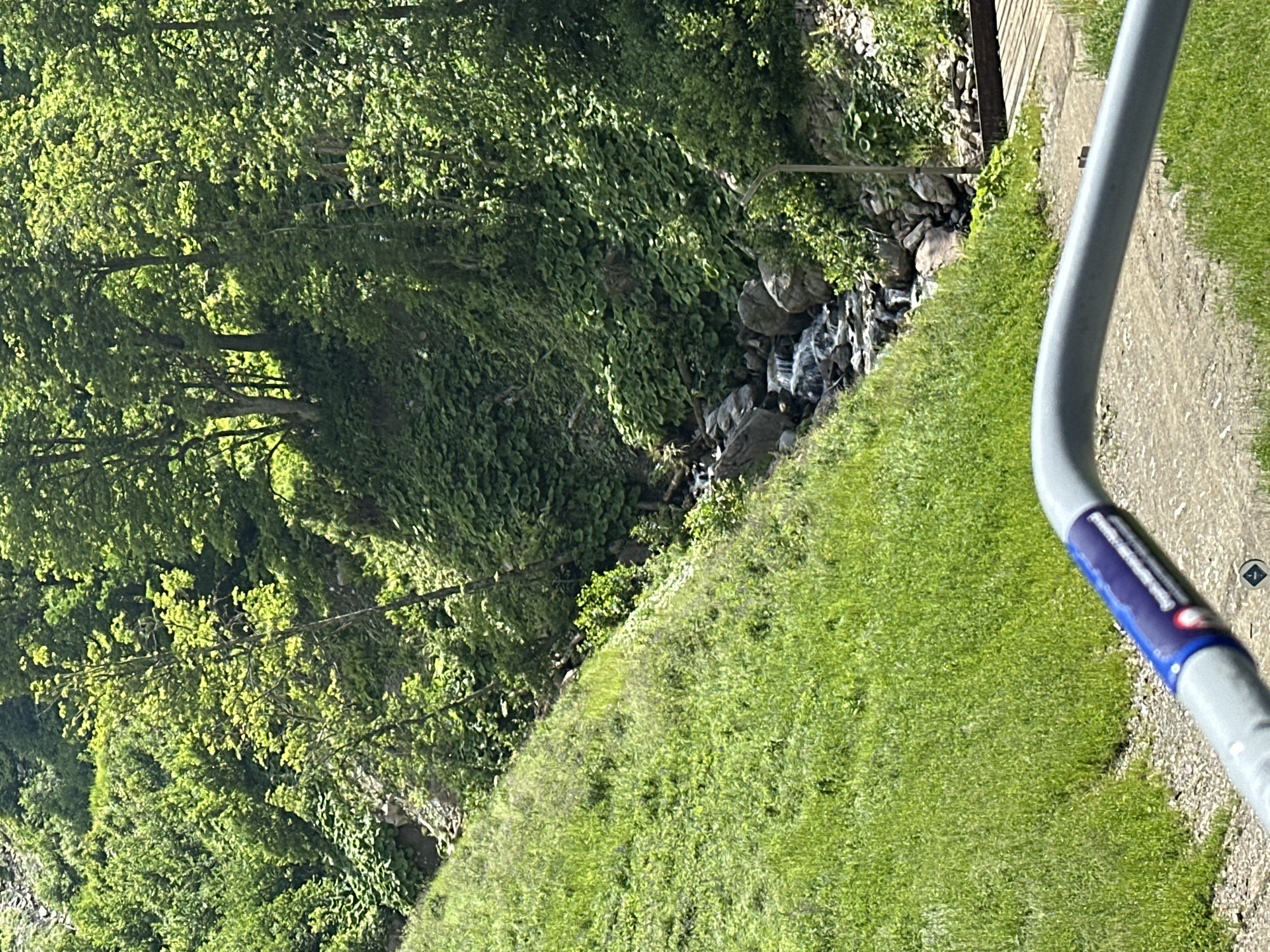 An outdoor, eye-level, medium shot captures a natural scene on a sunny day. The foreground features a grey railing with a blue and white sticker affixed to it. Beyond the railing, a path of gravel and dirt is visible, bordered by lush green grass on the left. The terrain slopes upwards towards the right.
Further into the frame, a small waterfall cascades over rocks into a stream. The stream is nestled within a dense forest area characterized by a variety of green foliage, including large, broad leaves and tall trees. The sunlight filters through the leaves, creating dappled patterns of light and shadow. The overall impression is one of a serene, natural landscape.
