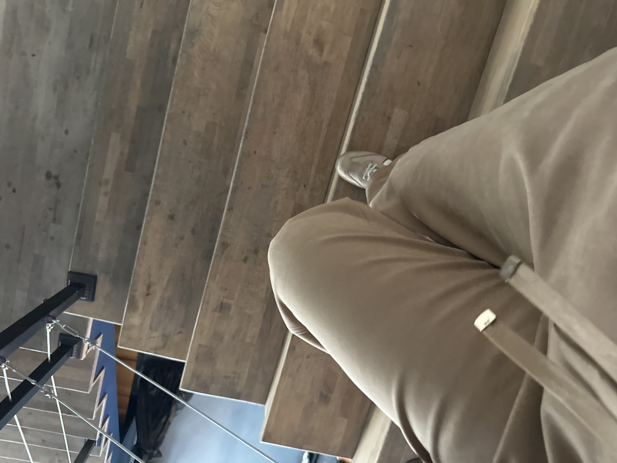 A first-person perspective photograph shows a person descending a wooden staircase. The person is wearing tan-colored pants and light-colored sneakers. The staircase has a black metal railing on the left side. Below the staircase, through the railing, a blue surface is visible. The location appears to be indoors, with potential evidence suggesting it could be in Marneuli, Georgia.