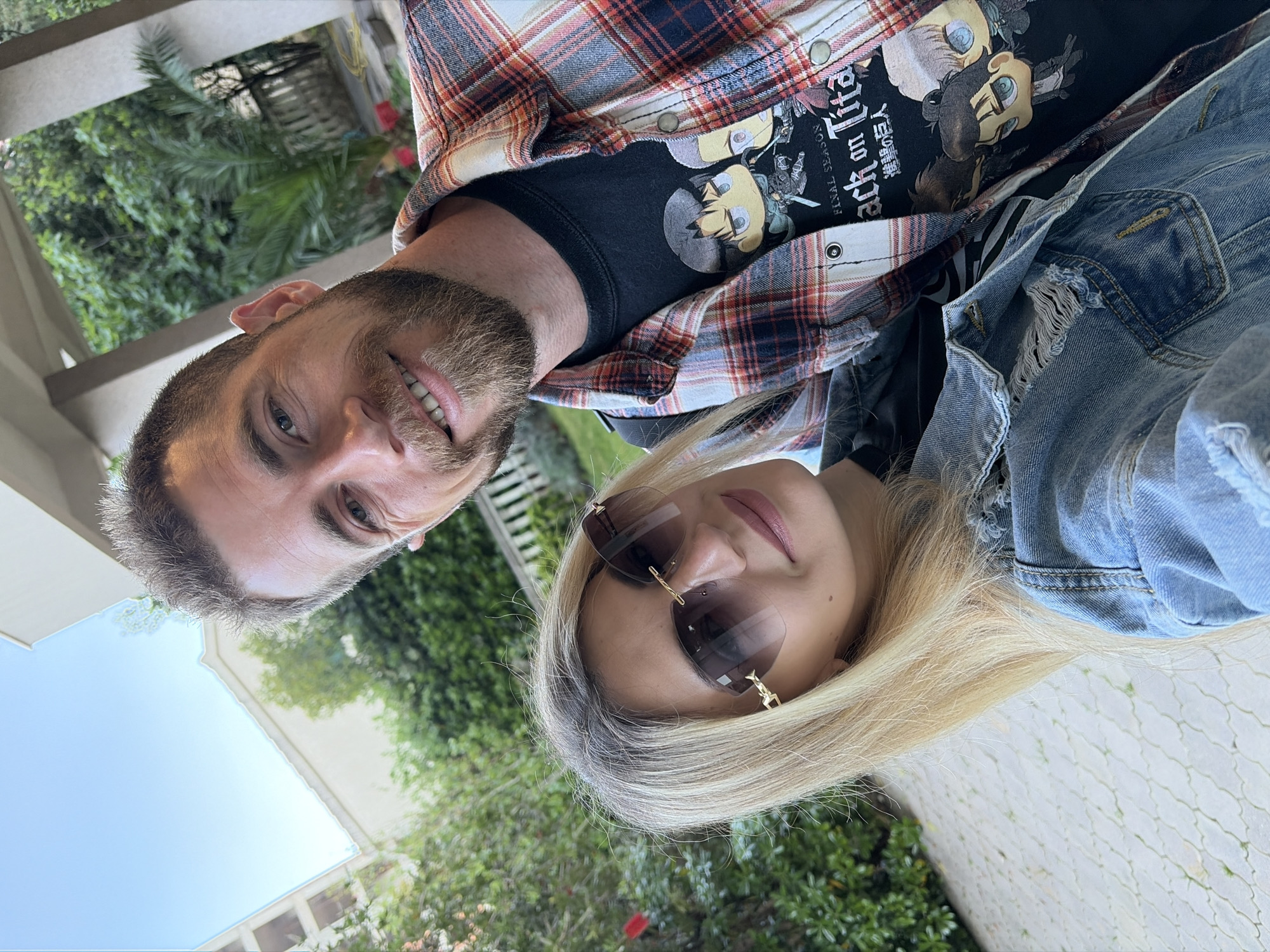 A selfie shows two individuals, a man and a woman, against an outdoor background with lush greenery and hints of architecture. The man, positioned to the left, has short, graying hair, a beard, and is smiling. He is wearing a red and white plaid shirt over a black t-shirt. The t-shirt features graphics, including stylized cartoon faces and Japanese text. The woman to the right has long, blonde hair and is wearing sunglasses and a denim jacket. The background includes foliage, glimpses of buildings, and paved ground. Without more specific information, it's not possible to identify the precise location in Budva, Montenegro. The image suggests an informal, possibly tourist-related snapshot.