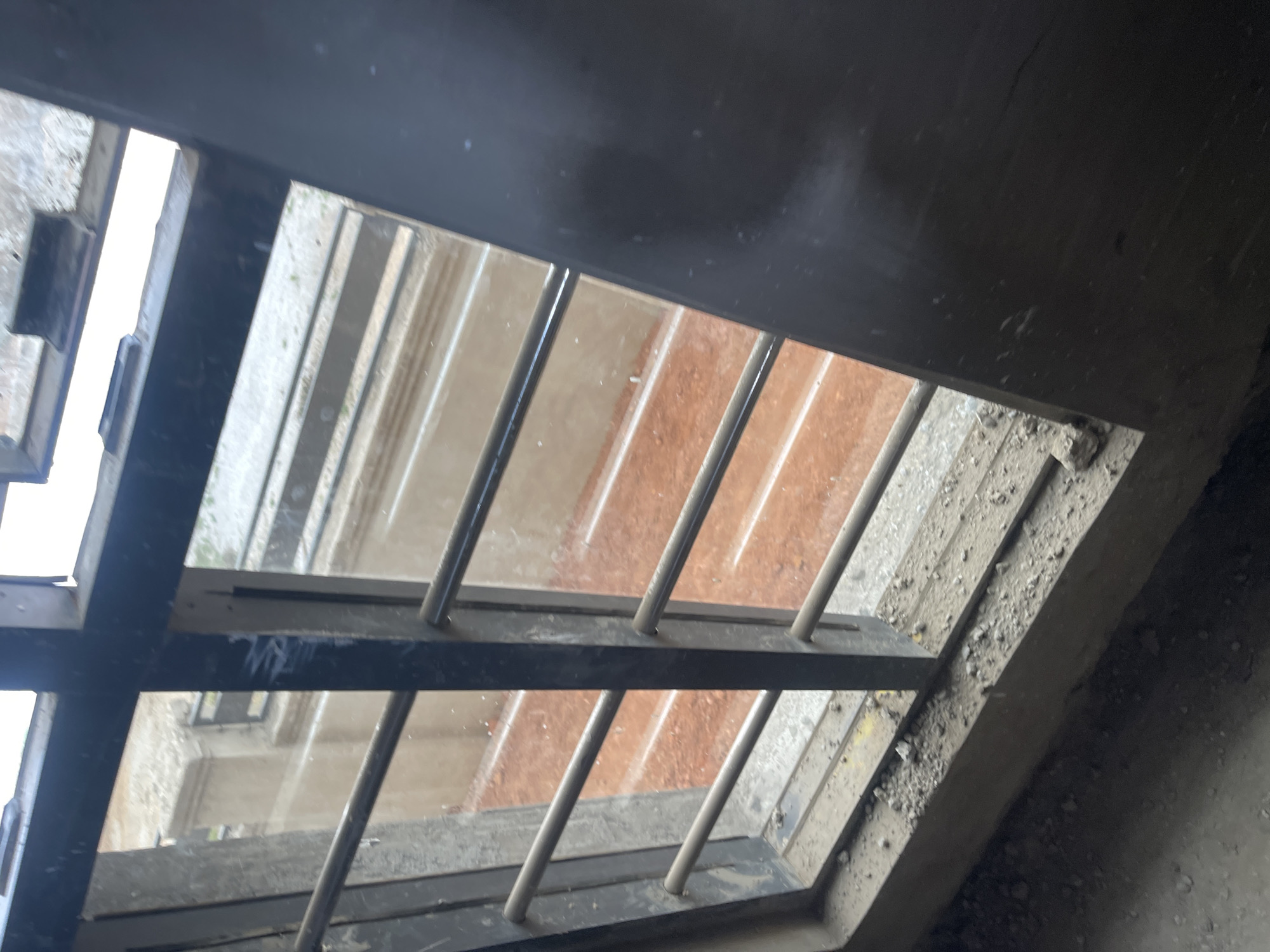 A close-up shot looking out through a barred window. The window frame and bars appear to be metallic and are dark in color. The walls around the window opening are rough and gray, possibly concrete or plaster. Through the window, one can see a building across the way, with architectural details suggesting multiple stories. The facade visible through the bars appears to have a combination of white or light-colored surfaces and sections that are terracotta or brick-red in color. The location is possibly a building in Angwan Sayawa, Nigeria, but this cannot be definitively confirmed from the image alone. There are no people visible in the scene.