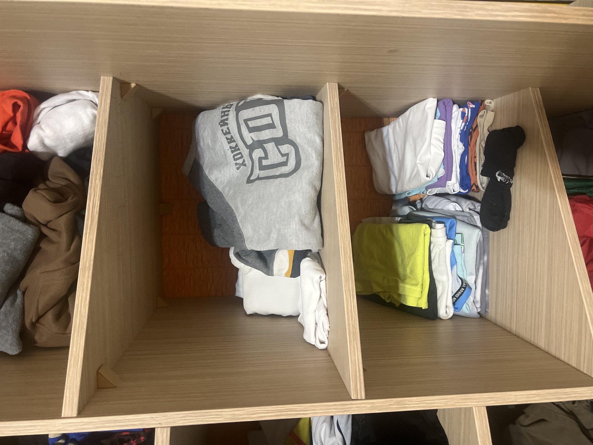 An indoor, eye-level, medium shot captures a closet with wooden shelves filled with folded clothing. The top shelf contains a brown hooded sweatshirt, a grey hooded sweatshirt, and a white cloth. The second shelf from the top holds a stack of white and grey t-shirts. The third shelf features a stack of t-shirts in various colors, including yellow, white, blue, and purple, with a pair of black socks in front of them. The bottom shelf is partially visible and appears to hold more clothing. The closet is well-lit, and the wooden shelves are clean and organized.