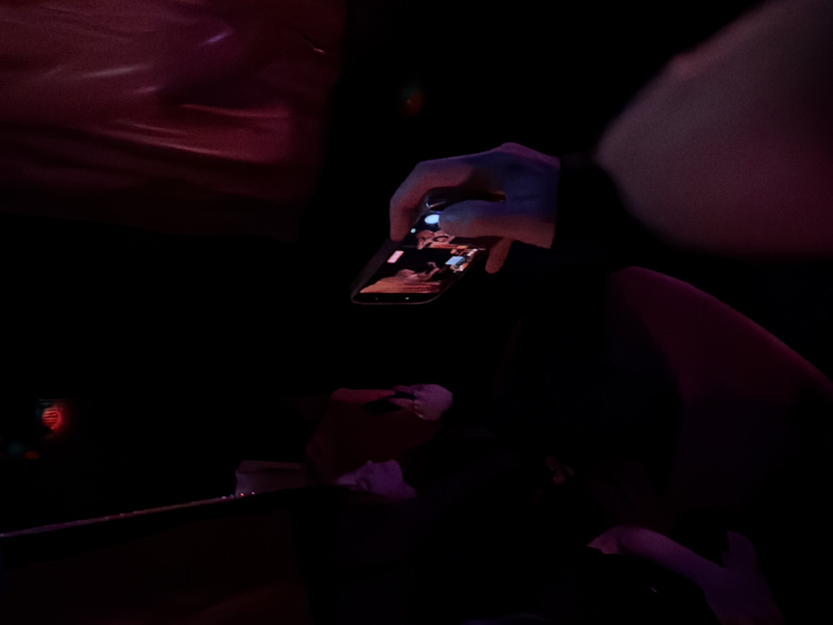 The image displays a dimly lit indoor environment dominated by red and purple ambient lighting. In the foreground, on the right side, a person's light-toned right hand holds a dark smartphone horizontally. The phone's screen is active, displaying a video recording or live camera feed featuring multiple figures in a brighter, possibly stage-lit, setting. A blue-white light emanates from a specific point on the phone's screen. To the left, a person's profile is partially visible in silhouette, illuminated by purple light, observing the scene towards the right. Their left hand is also partially visible. The upper left background features a circular object emitting a red glow, while the upper right background shows indistinct dark or reddish material. Additional human legs are visible in the lower left, also illuminated by purple light. The specific location within Paris, France, is not identifiable from the visual information provided; however, the scene suggests a nightlife or entertainment venue during an event.