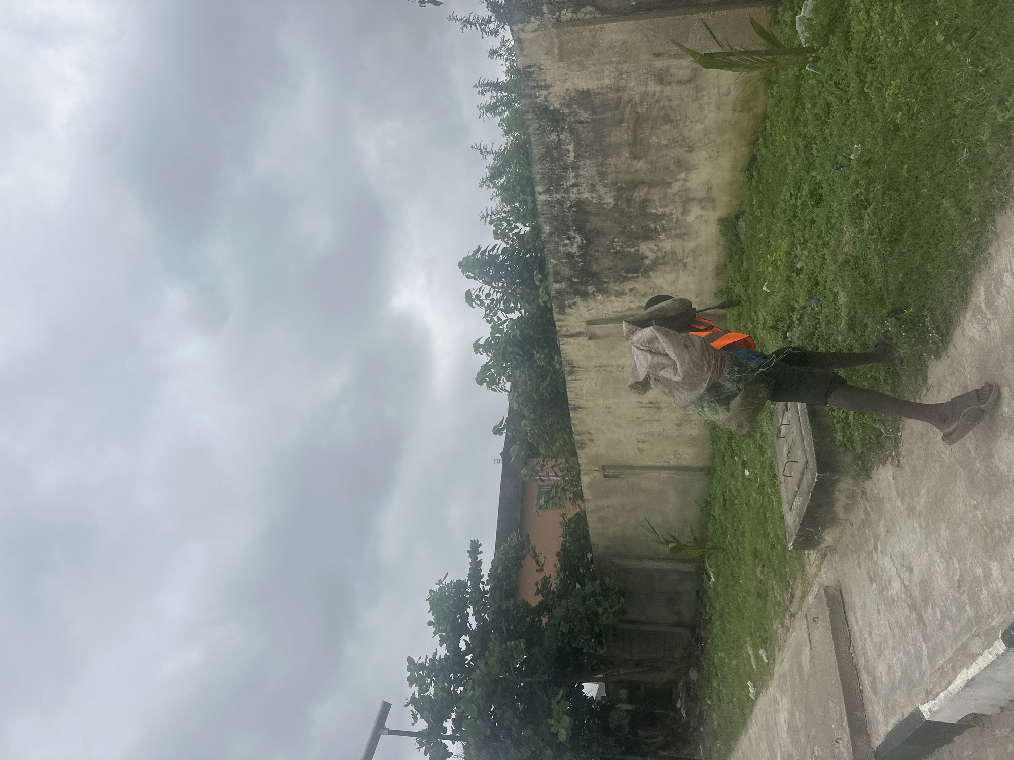 A person, dressed in dark shorts and an orange vest over a dark garment, carries a large sack of material over their left shoulder. Their right leg is extended forward, mid-stride, and they are wearing dark sandals. The person is walking along a concrete pathway adjacent to a grassy area. In the background, a weathered concrete wall runs horizontally, with lush green foliage and trees growing along its top and behind it. A building with an orange facade is visible behind the trees. The sky is overcast with heavy grey clouds. The scene appears to be in an urban or semi-urban environment in Badagry, Nigeria, possibly near a residential or commercial area given the building and wall.