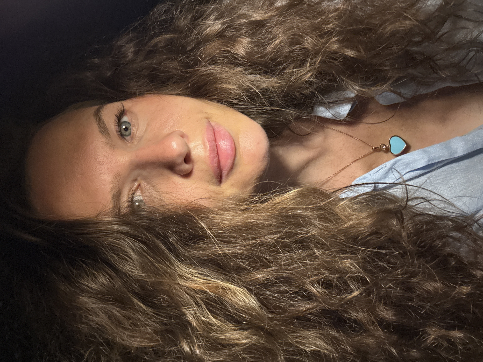 A close-up, medium shot captures a woman with long, curly brown hair and light-colored eyes. She is looking towards the camera, and the lighting casts a warm glow on her face, highlighting her nose and lips. She is wearing a light blue collared shirt, open at the neck, revealing a rose gold necklace with a heart-shaped pendant of a bright blue stone. The background is dark and out of focus. The image was taken in Dubai, United Arab Emirates.