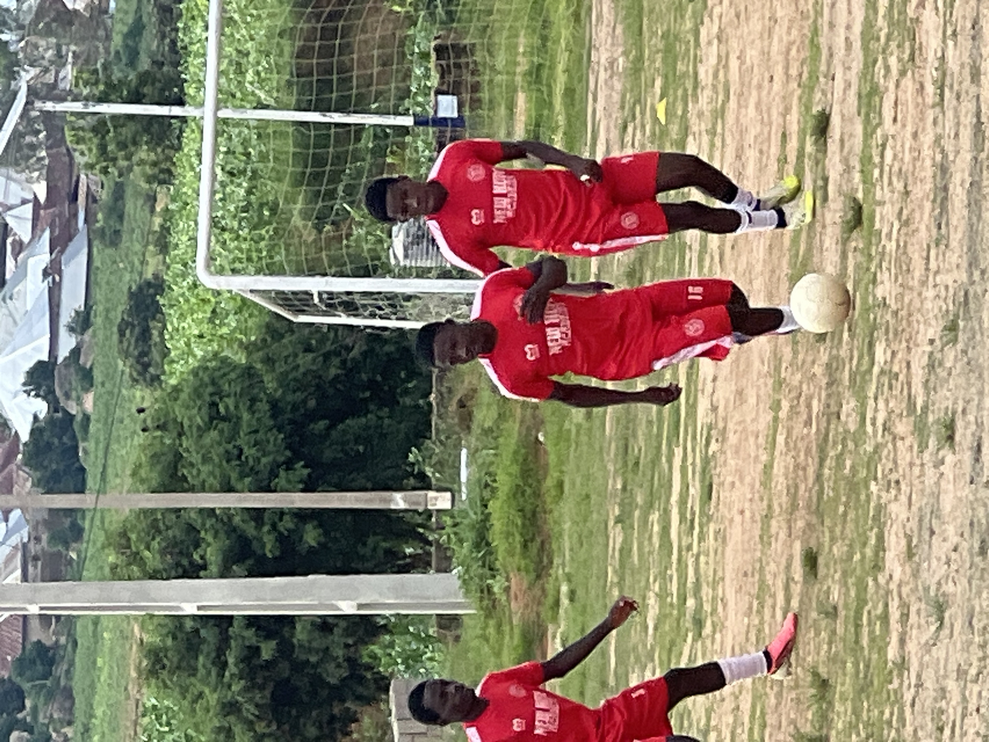 Three young men in red soccer uniforms are on a dirt field with sparse grass, playing soccer. Two are actively engaged with a soccer ball, while the third is further back and appears to be running. The background shows a soccer goal with a net, trees, and buildings in the distance, suggesting an outdoor sports facility. The sky is overcast. The scene is consistent with a soccer practice or casual game in a developing area. This image could be from the city of Jos, Nigeria, given the visual cues of the environment and the people depicted.