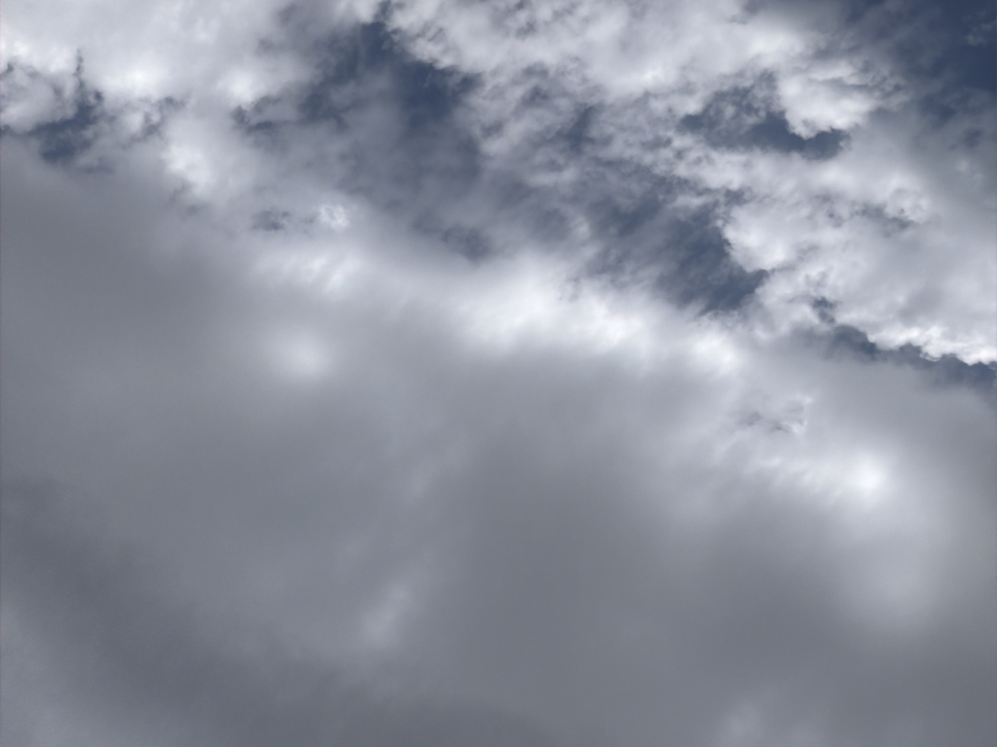 This is a vertical, medium-angle shot of the sky above Oxentea, Moldova. The sky is filled with a mix of grey and white clouds. On the left side of the frame, the clouds are dense and uniform grey, obscuring any view of the blue sky. As the image moves to the right, the clouds become thinner and more fragmented, revealing patches of dark blue sky in between. These white and grey clouds are textured, with some appearing wispy and others more voluminous. A faint, diffused light source, possibly the sun, is visible through a thin patch of cloud towards the upper right quadrant of the image. There are no people, animals, or man-made structures visible in this image. The scene appears to be an outdoor, aerial view.