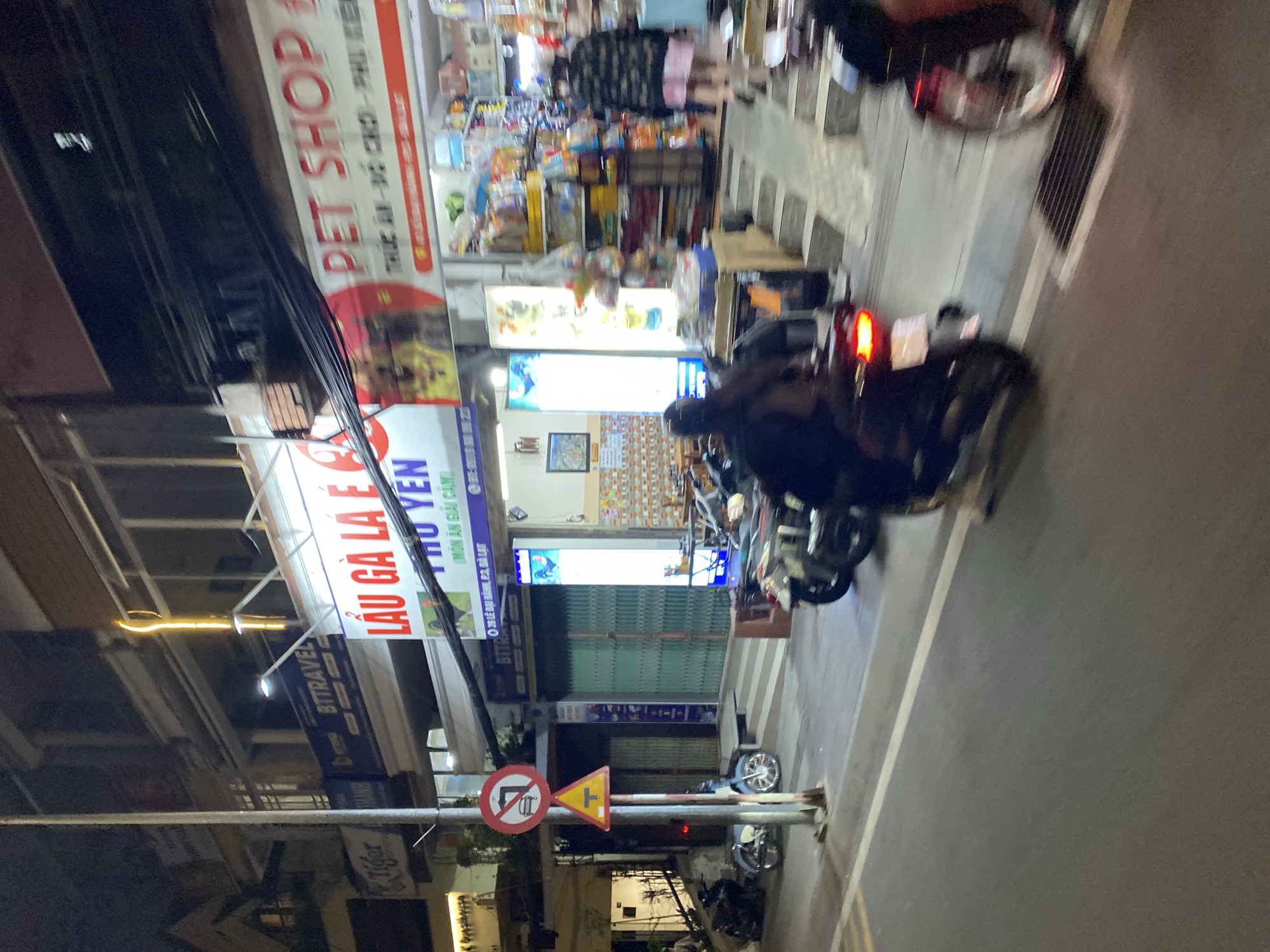 The image captures a bustling street scene at night in Da Lat, Vietnam, characterized by bright shop lights and traffic. The perspective is tilted, showing the street on the right and buildings on the left.
The street is illuminated by artificial light sources, including overhead streetlights and brightly lit shopfronts. Several people are present; some are seen on motorcycles, with one motorcycle in the foreground showing a red brake light, suggesting it is either stopped or moving slowly. Another individual appears to be walking on the pavement. The street itself is paved with a white line marking its edge.
Numerous shops line the street. Prominently visible is a large, illuminated sign for "LẨU GÀ LÀ É C THO YEN (MÓN ĂN GIẢI CẢM)", which translates to a chicken hotpot restaurant, likely named "Tho Yen", specializing in dishes for cold relief. Below this, an address is partly visible: "39 LÊ ĐẠI HÀNH, P.2, ĐÀ LẠT", confirming the specific location in Da Lat. Another sign for a "PET SHOP" is visible further down the street, advertising "THỨC ĂN - ĐỒ CHƠI - PHỤ KIỆN" (Food - Toys - Accessories). A sign for "BTTRAVEL" indicates a travel agency, and a "Tiger" logo is also discernible on a banner.
On a pole in the middle ground, two distinct traffic signs are visible: a circular sign with a red border and a line through a left-turning arrow, indicating "No Left Turn" or "No U-Turn", and a yellow triangular warning sign, possibly indicating a T-junction or other road hazard. Overhead, a tangle of electrical wires is a common sight in urban Vietnamese landscapes. The overall impression is one of an active urban night, full of commerce and movement.