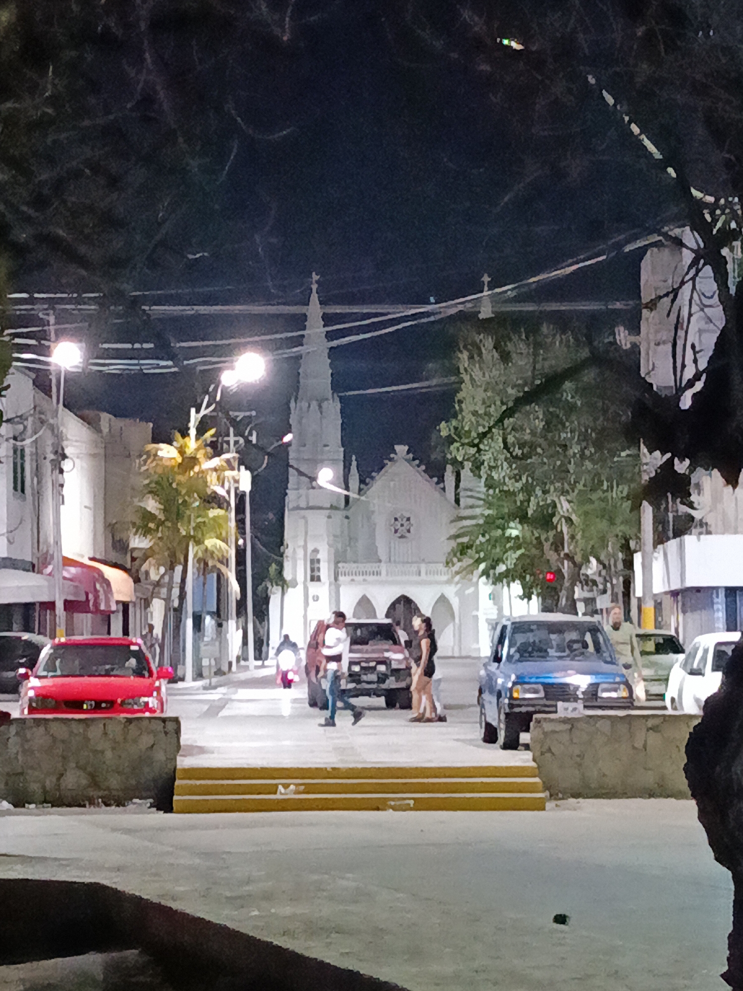 The image shows a street scene at night in Juan Griego, Venezuela. In the background, a white church with a tall spire is visible. Several cars, including a red one on the left and a blue one on the right, are parked along the street. People are standing and walking near the cars and on the sidewalk. Streetlights illuminate the scene. A small staircase with yellow edges leads from the foreground to the street. Trees are partially visible on the sides of the frame, obscuring the view. Power lines run above the street.