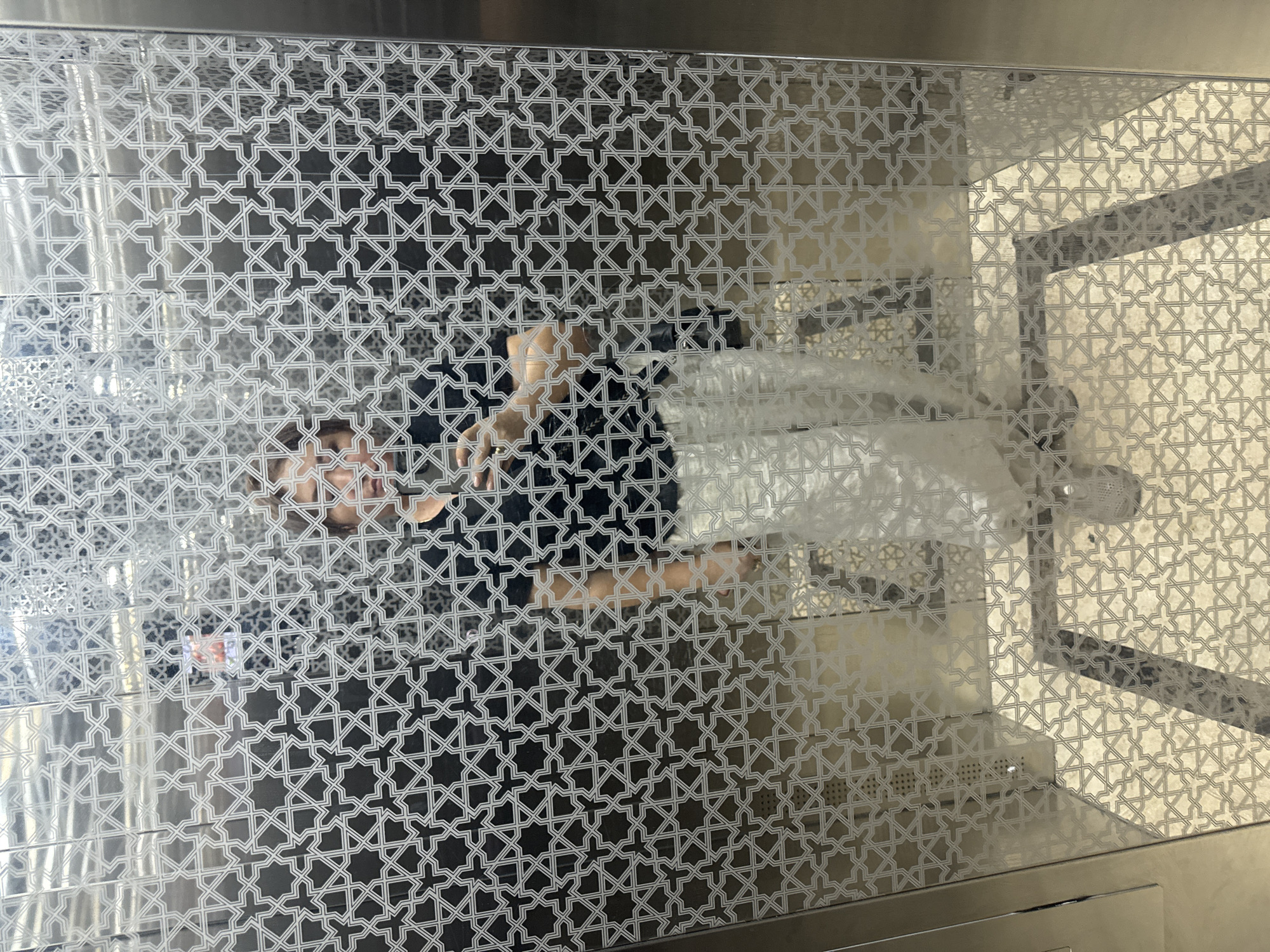 A woman is reflected in a patterned mirrored wall, holding a phone and taking a selfie. She is wearing a black t-shirt and white pants. The elevator interior features a mirrored wall with a repeating geometric pattern and a patterned floor. The scene is brightly lit, and the woman's face is partially obscured by the pattern.
