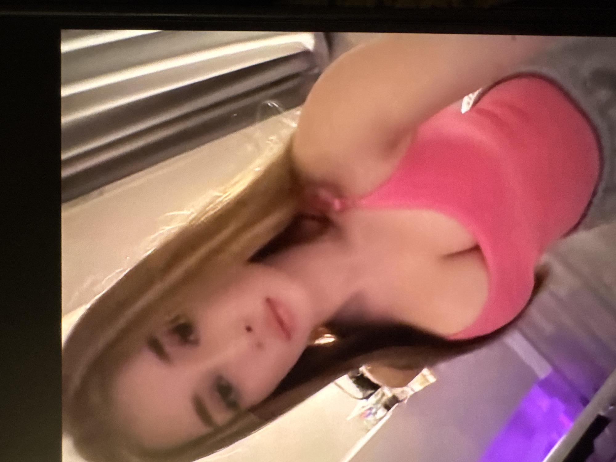 A young woman with fair skin and light brown hair is posing for a selfie. She's wearing a pink tank top and a gray sleeveless shirt. The background is blurry, but suggests a room with blinds. There is no way to determine if the location is in Yerevan, Armenia.