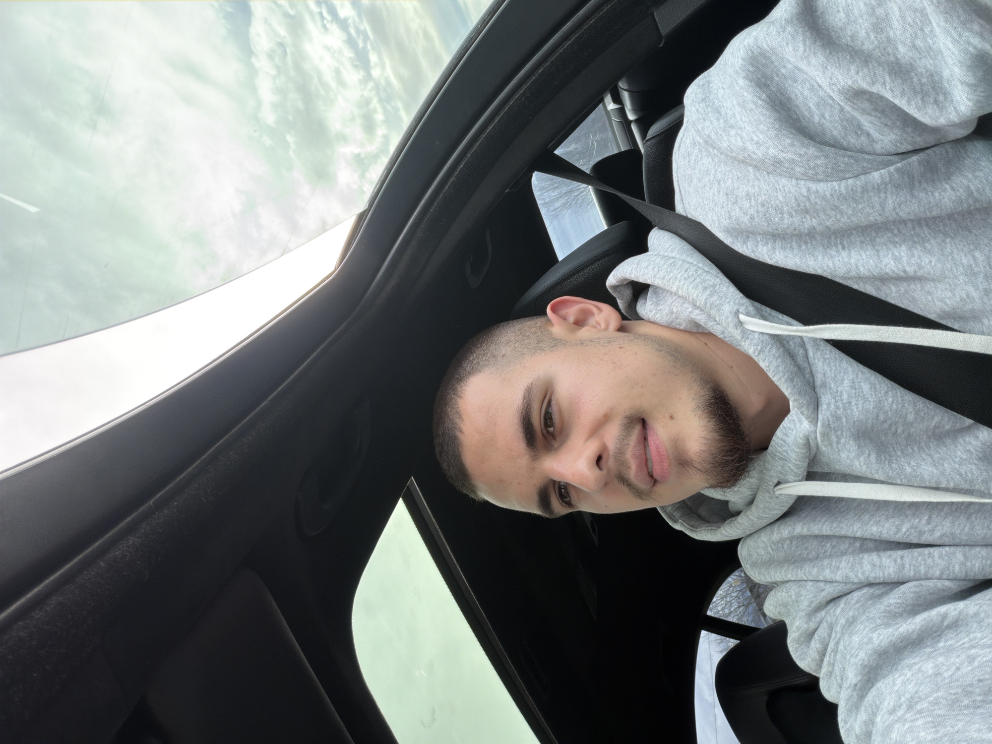 A young man sits in the driver's seat of a car with a sunroof. He is wearing a grey hooded sweatshirt and a black seatbelt. He looks toward the camera with a slight smile. The sunroof offers a view of an overcast sky. The car's interior is black. The location in Plovdiv, Bulgaria, cannot be determined based on the available visual information.