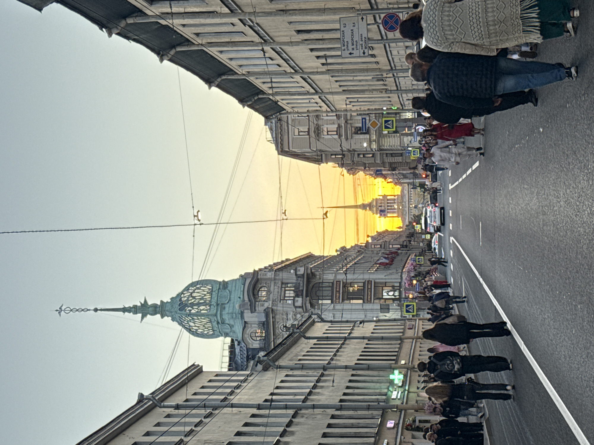 The image depicts a street scene in Saint Petersburg, Russia, likely Nevsky Prospekt, during sunset. A wide asphalt road runs through the center of the frame, lined with imposing, multi-story buildings on both sides. The architecture features classical European elements with numerous windows and decorative facades. A distinctive green, ornate dome with a spire tops a building on the left, and a similar spire is visible in the distance at the end of the street, silhouetted against the vibrant orange sunset sky.
Numerous people are walking on the sidewalks and crossing the street, dressed in casual attire. Some are walking in groups, while others are alone. Several cars are visible on the road, and traffic lights and street signs are present. The atmosphere appears to be lively, with pedestrians engaged in their daily activities. The overall impression is that of a bustling urban environment captured during a beautiful evening sunset.