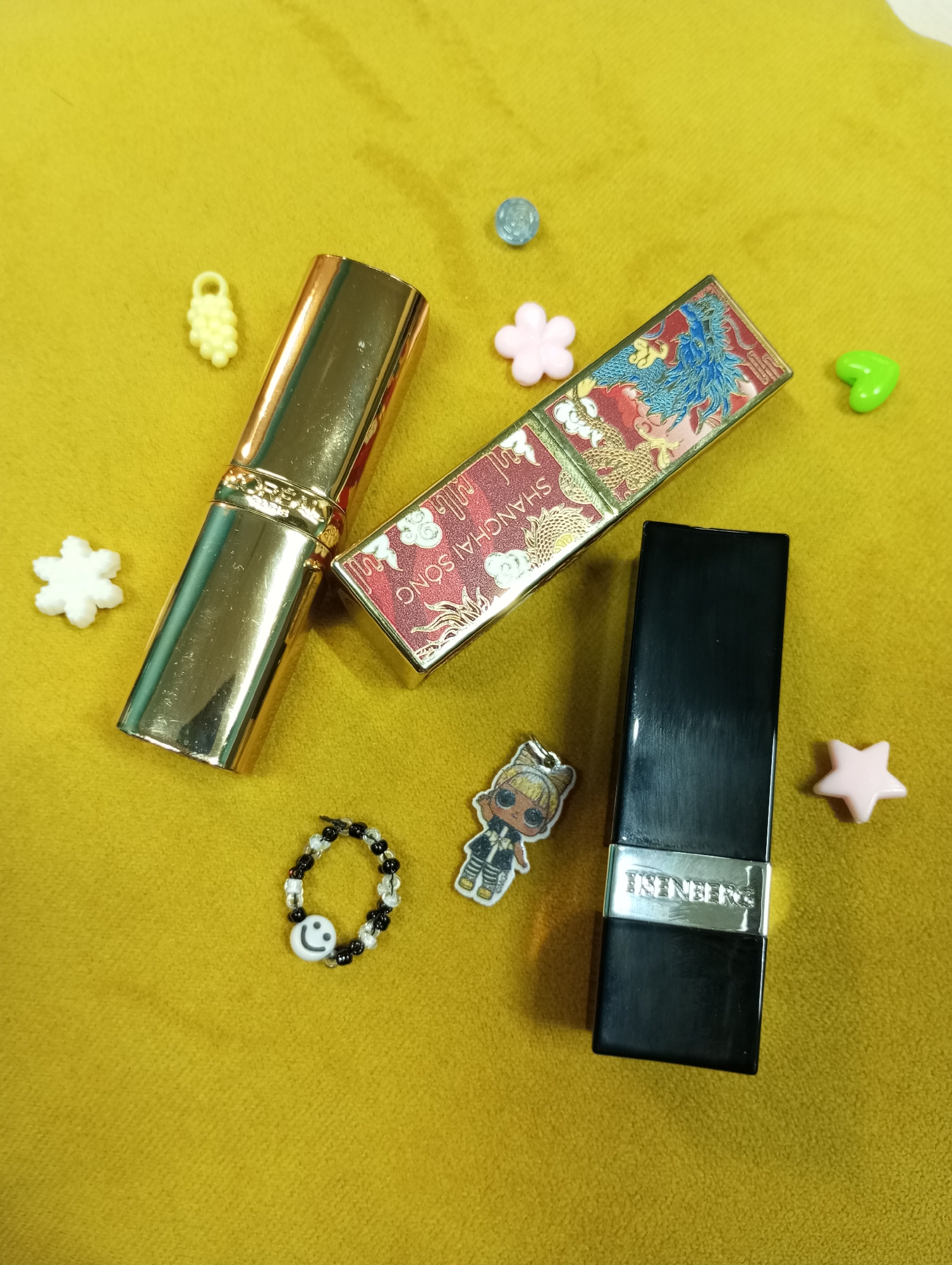The image depicts three lipsticks and several small trinkets on a yellow surface. A gold-colored lipstick is to the left, a red lipstick with a dragon design is in the center, and a black lipstick is to the right. Around the lipsticks are a small smiley face ring, a doll sticker, and several plastic flower and star-shaped beads. No details indicate the city of Pervomaysky, Russia.