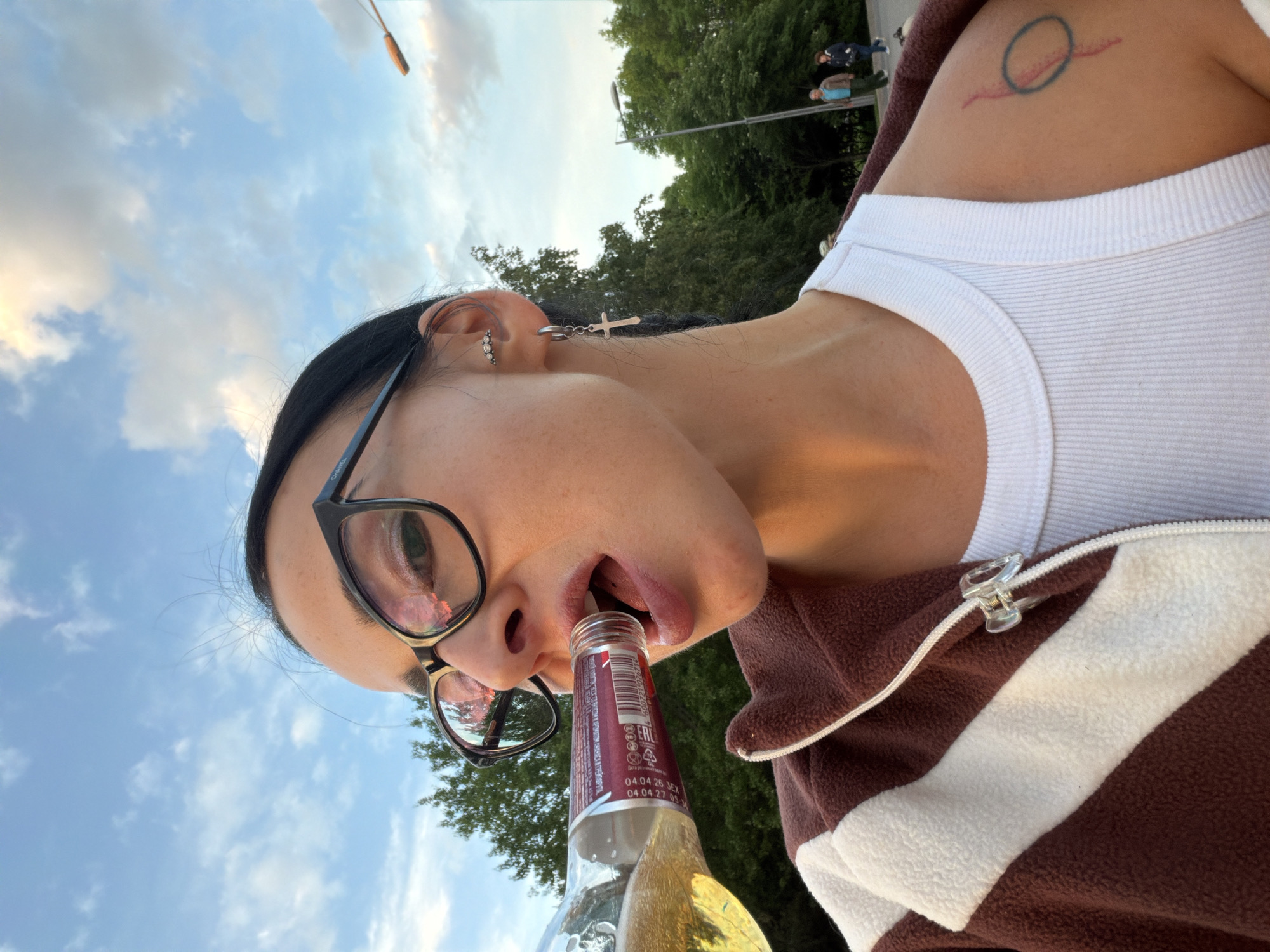 An upward-angled, first-person perspective photograph captures a person drinking from a bottle outdoors in Saint Petersburg, Russia. The individual, seen from the nose downwards and to the side, is wearing black-rimmed glasses with "CHANEL" faintly visible on the left temple arm, a white ribbed tank top, and a brown and white striped fleece jacket. They have multiple earrings, including a small hoop with stones and a dangling cross, and dark hair pulled back. A distinct tattoo featuring a blue circle with a red line underneath is visible on their upper left shoulder.
The person is holding a clear glass bottle with a red and white label to their mouth, appearing to drink a light-colored, bubbly liquid, likely a beverage such as beer or cider. Text on the bottle label includes "04.04.26," "04.04.27," "EAC," and a recycling symbol, along with other unidentifiable Cyrillic script.
The background primarily features a clear blue sky with scattered white clouds, suggesting good weather. Green trees are visible in the middle distance, and in the far background on the right, blurred figures of two people and a utility pole or street light indicate an outdoor public setting. The soft, bright lighting points to late afternoon or early evening. The overall composition conveys a casual and candid moment.