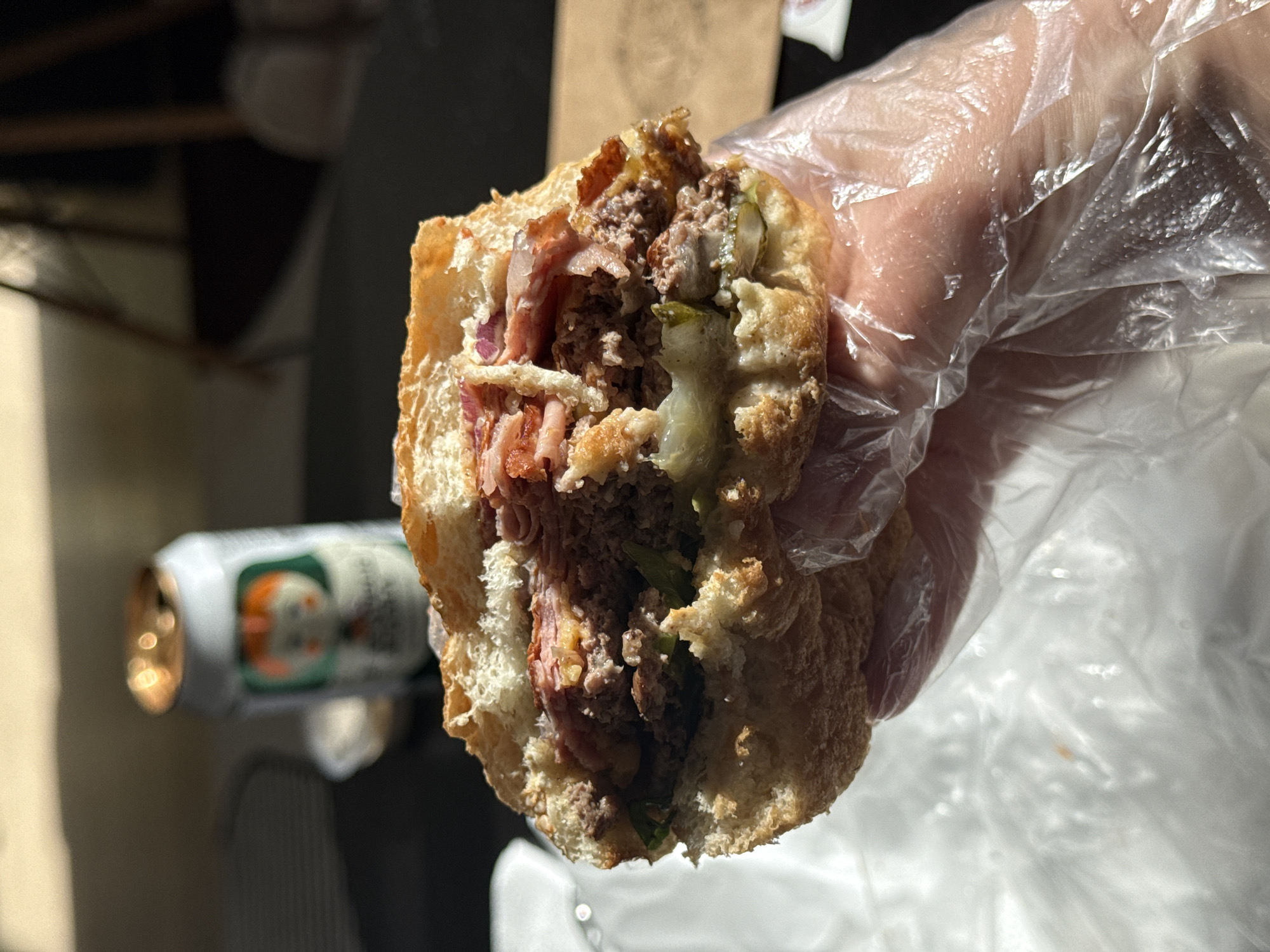 The image shows a half-eaten sandwich held in a gloved hand. The sandwich appears to be a meat-filled roll, possibly a hoagie or sub, with visible ingredients including ground meat, sliced meat (possibly ham or prosciutto), pickles, and possibly red onion. The bread has a browned crust. In the background, out of focus, is a can of soda and what appears to be a paper bag. The location, Vozdovac Urban Municipality, Serbia, cannot be definitively determined from the image.