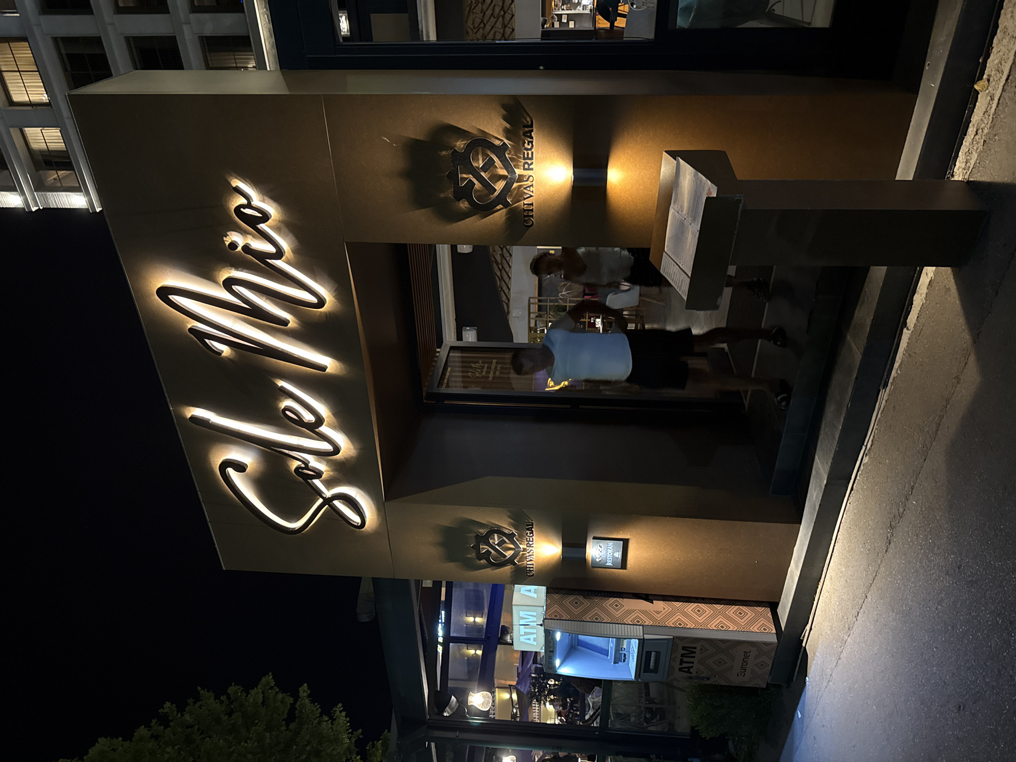 The image shows the entrance to a restaurant named "Sole Mio" at night, located in Budva, Montenegro. The restaurant has a prominent illuminated sign with the name "Sole Mio" in elegant script. To the left of the entrance, there is an ATM machine branded "Euronet". On the wall next to the entrance, there is a logo and text that reads "CHIVAS REGAL", illuminated by a spotlight. Further down the wall, a smaller sign displays the word "RESTORAN". Two people, a man and a woman, are walking into the restaurant through the open doorway. The man is wearing a white t-shirt and dark shorts, and the woman is wearing a light-colored top and dark shorts. The street in front of the restaurant is paved and dimly lit. A modern building with illuminated windows is visible in the background, to the right of the restaurant.