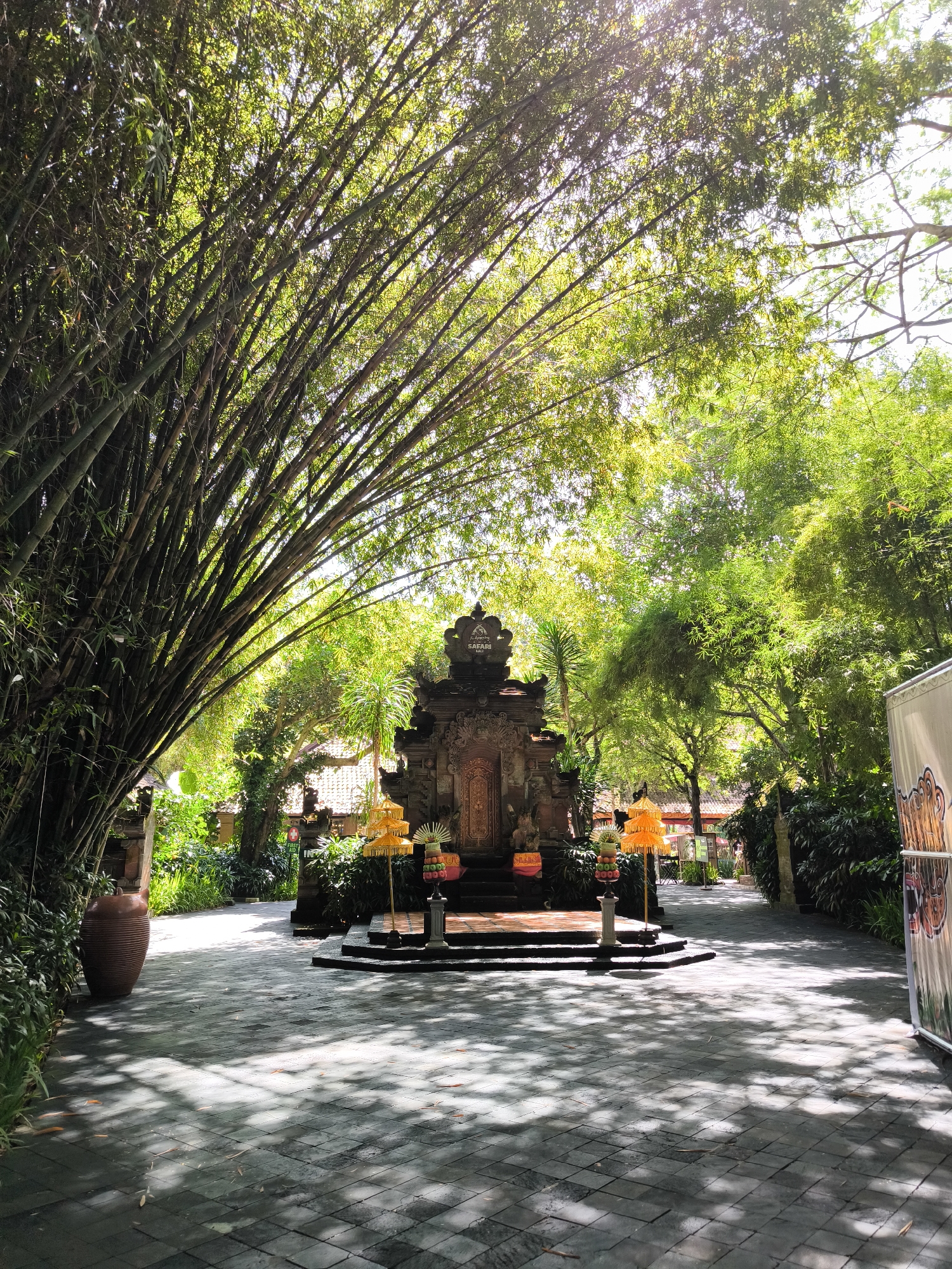 An outdoor shot depicts a stone pathway in front of a Balinese-style temple. The pathway is paved with dark grey square tiles that are arranged in a diagonal pattern and are partially covered in shadow. To the left of the pathway, a large, dark brown clay pot stands next to lush green foliage. The temple itself is ornate, with dark stone carvings and a tiered roof. It is flanked by two tall, golden umbrellas and a decorative display of colorful fruit. A pathway leads away from the temple, surrounded by dense green trees and foliage. The entire scene is bathed in the dappled sunlight that filters through the overhanging bamboo stalks, creating a dramatic interplay of light and shadow. The lighting suggests it is daytime.