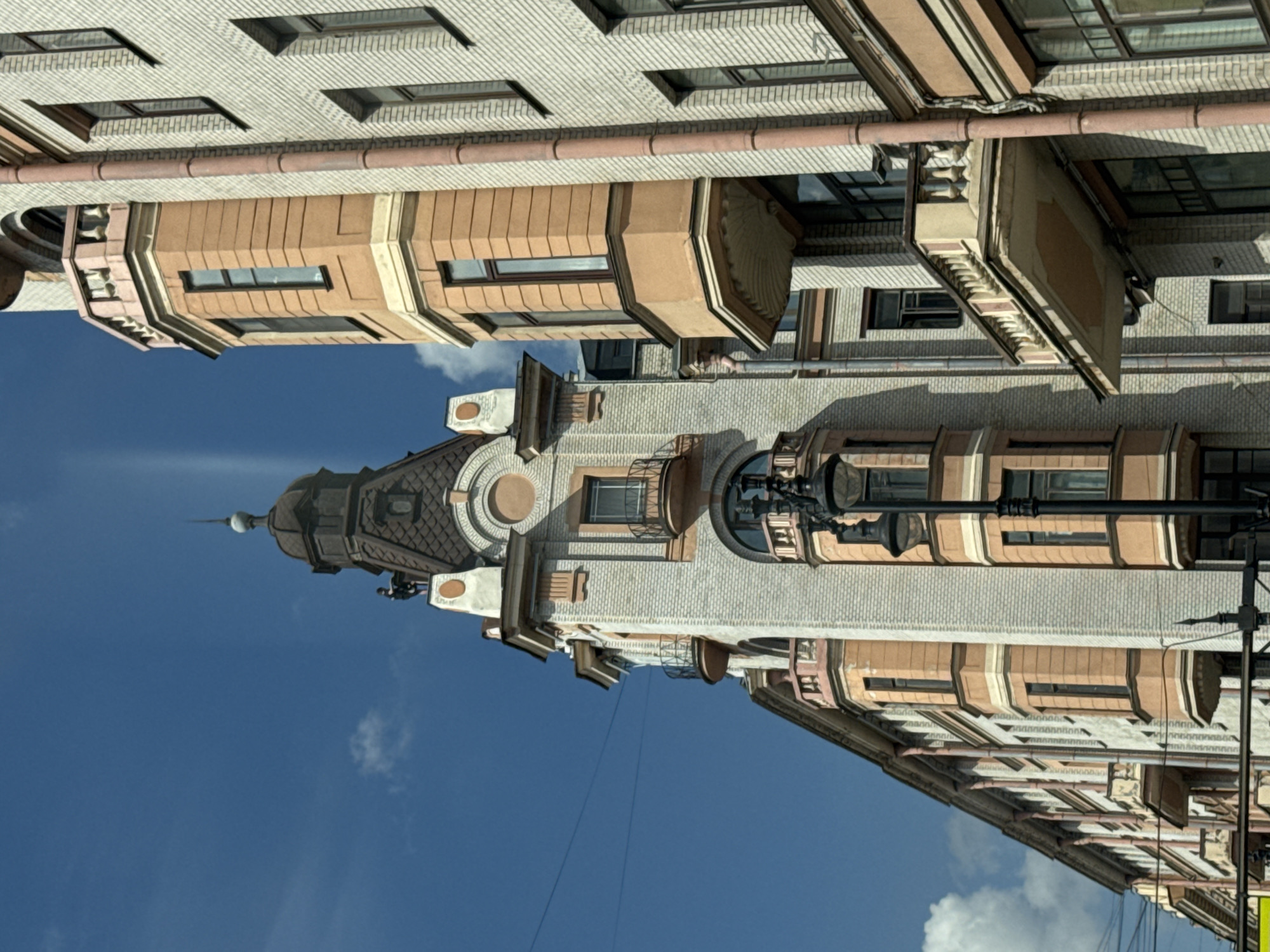 The image presents an upward view of an ornate, multi-story historic building in Saint Petersburg, Russia, set against a clear, bright blue sky dotted with scattered white clouds. The building's main facades are constructed from light-colored, possibly cream or off-white, brickwork with a distinctive patterned texture.
The architecture is rich with detail, featuring projecting bay windows that are both rounded and polygonal, accentuated by contrasting yellowish-beige or tan-colored decorative elements. These lighter shades also frame the numerous rectangular windows and form horizontal bands across the facade. Dark reddish-brown drainage pipes are visible running along the building.
A prominent feature is a tall, elaborate corner tower or turret, culminating in a pointed spire. This tower showcases intricate roofing, likely made of dark tiles, and includes smaller windows and decorative accents. Balconies with ornate railings and decorative cornices add to the building's complex design. On the right side, a black, traditional lamppost extends vertically, partially visible.
The scene depicts a sunny daytime setting, suggesting late morning or early afternoon due to the bright, direct light. The weather is fair with minimal cloud cover. No people are visible within the frame, and there is no discernible text on the building or any other objects. The overall impression is one of grand, turn-of-the-century European urban architecture.